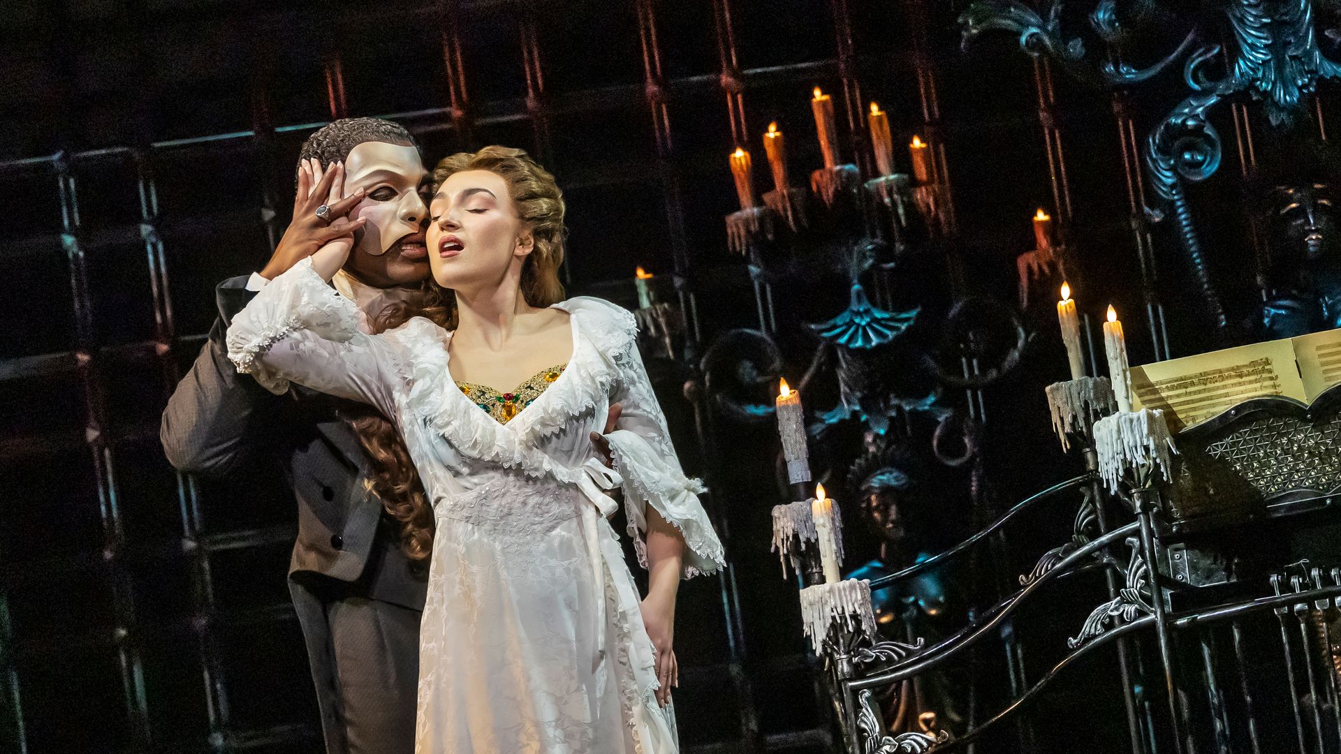 On a gothic stage, a masked man in a dark suit cups a woman's face as she tilts back in a white lace gown; candlelit candelabras and sheet music flank an ornate iron desk.
