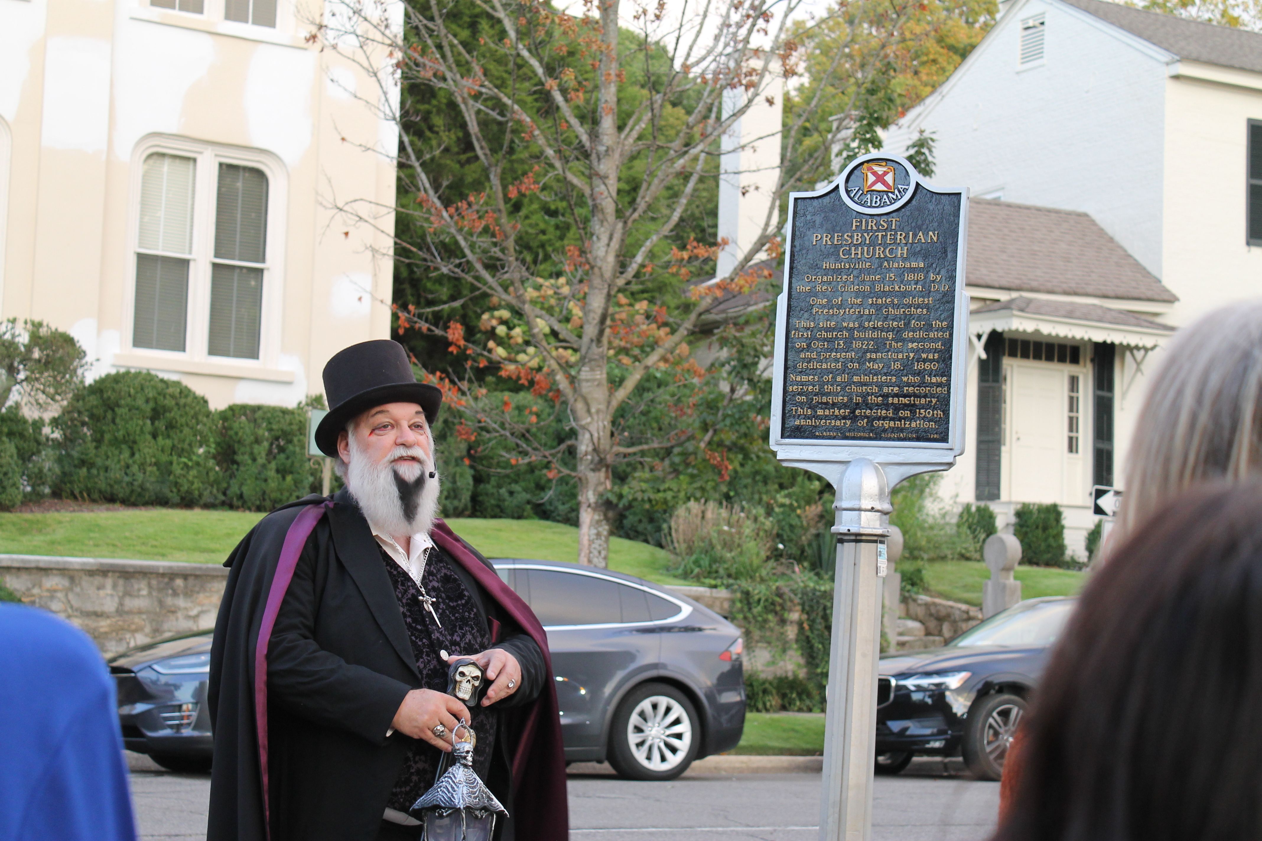 A man in a black top hat, black and purple cape, and white beard stands next to a historical marker for First Presbyterian Church in Huntsville, Alabama, speaking to a small crowd.