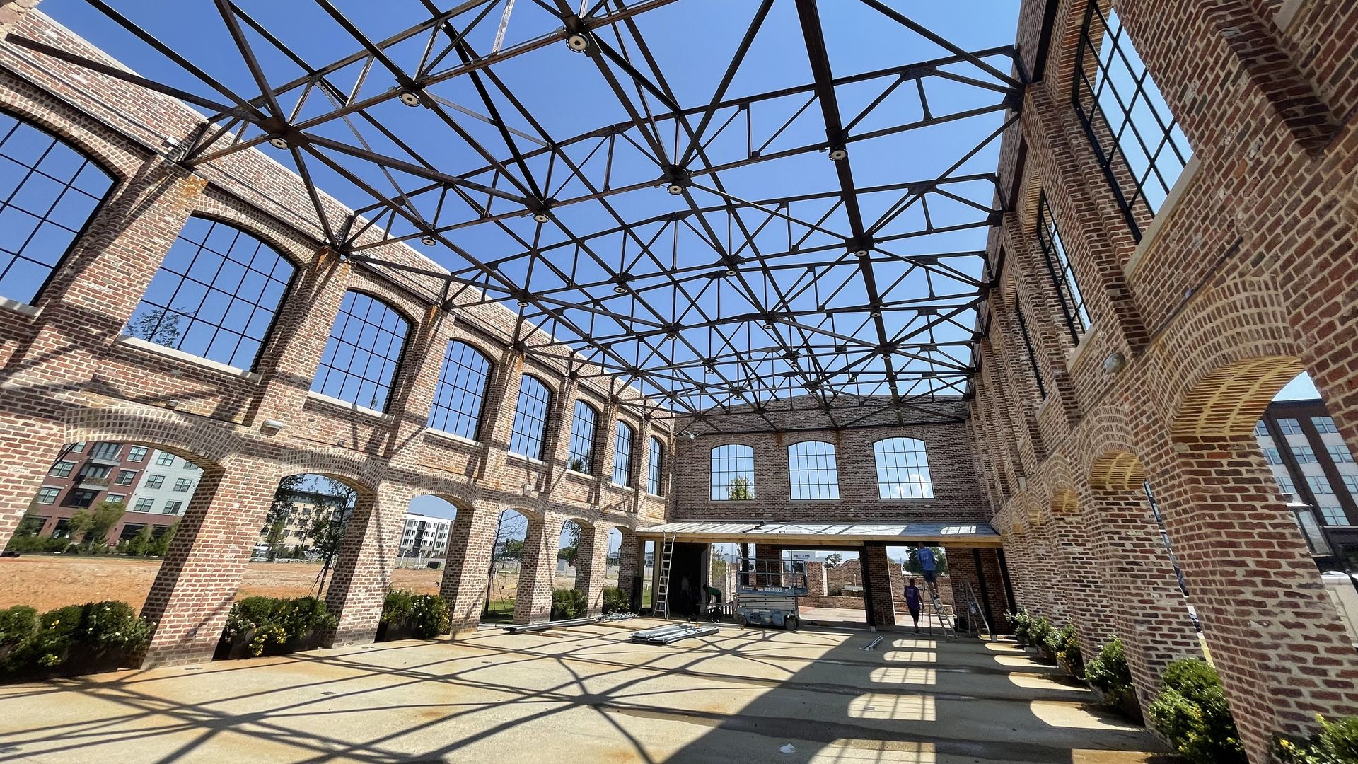 Open brick building with large arched windows and exposed black metal roof beams under clear blue sky. Shadows and a few people near scaffolding inside.
