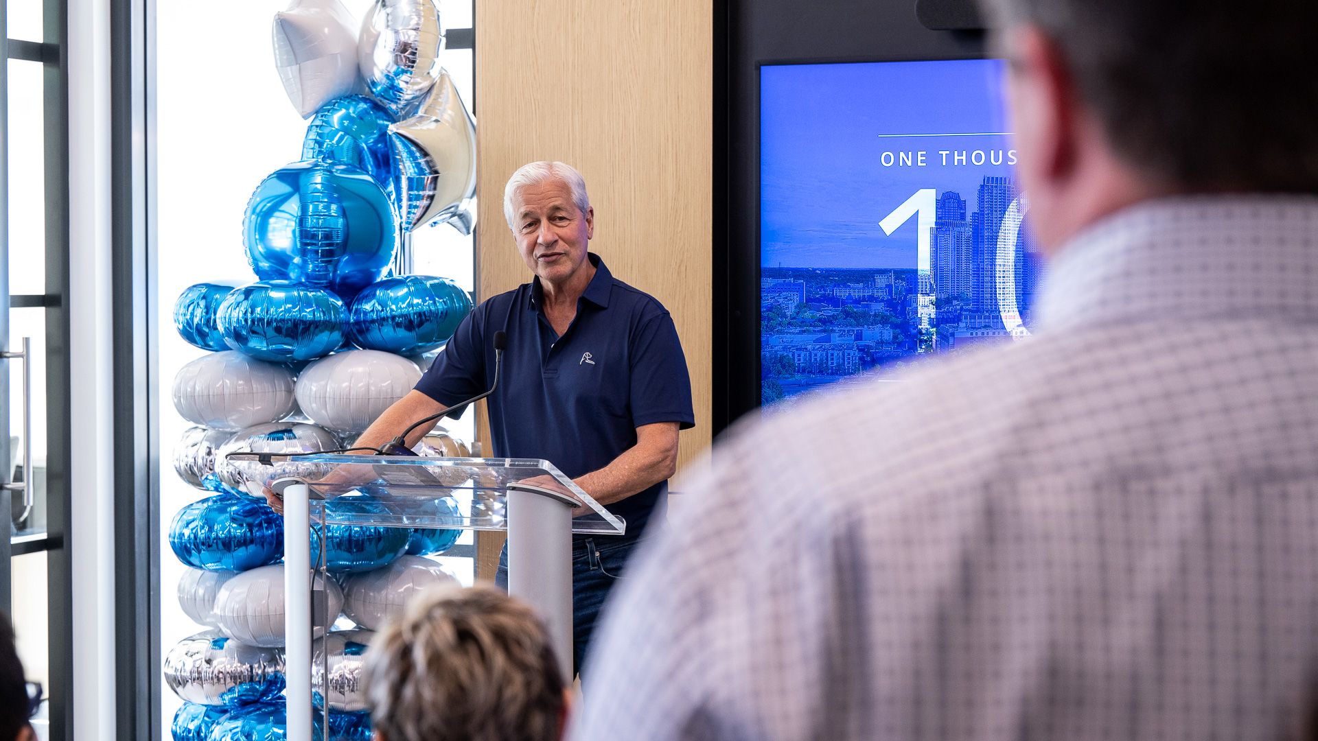 Older man with white hair speaks at a clear podium; navy polo. Behind him a blue cityscape screen; to his left stacked blue and silver balloons; audience in foreground.