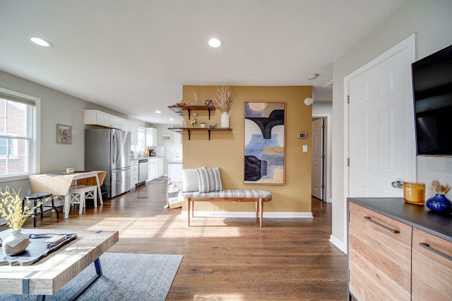 Bright open living-kitchen space with white cabinets, a stainless fridge, a mustard accent wall, floating shelves, abstract art, a cushioned bench, and warm wood floors.