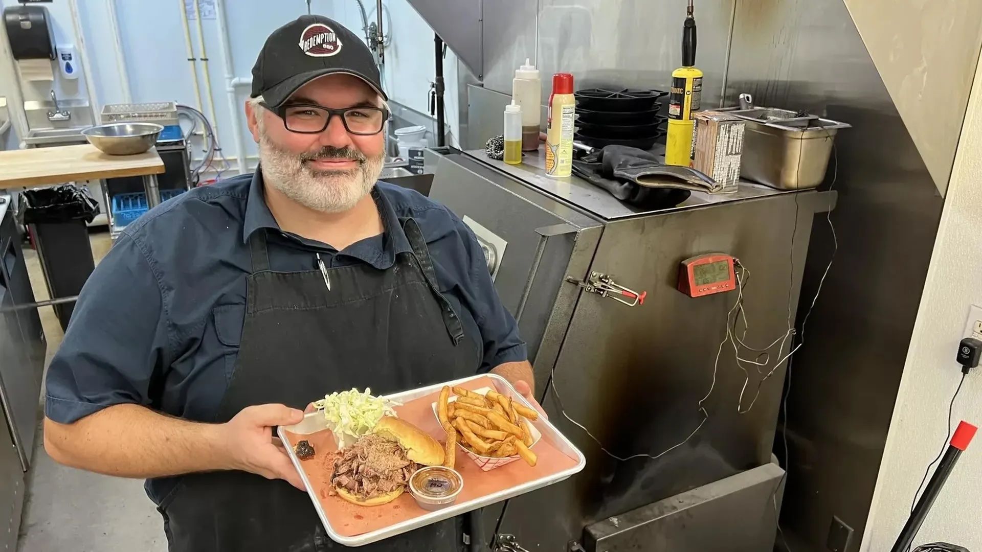 Smiling man in a black cap and apron holds a tray with a sandwich, fries, coleslaw, and sauce in a stainless steel kitchen with a large smoker in the background.