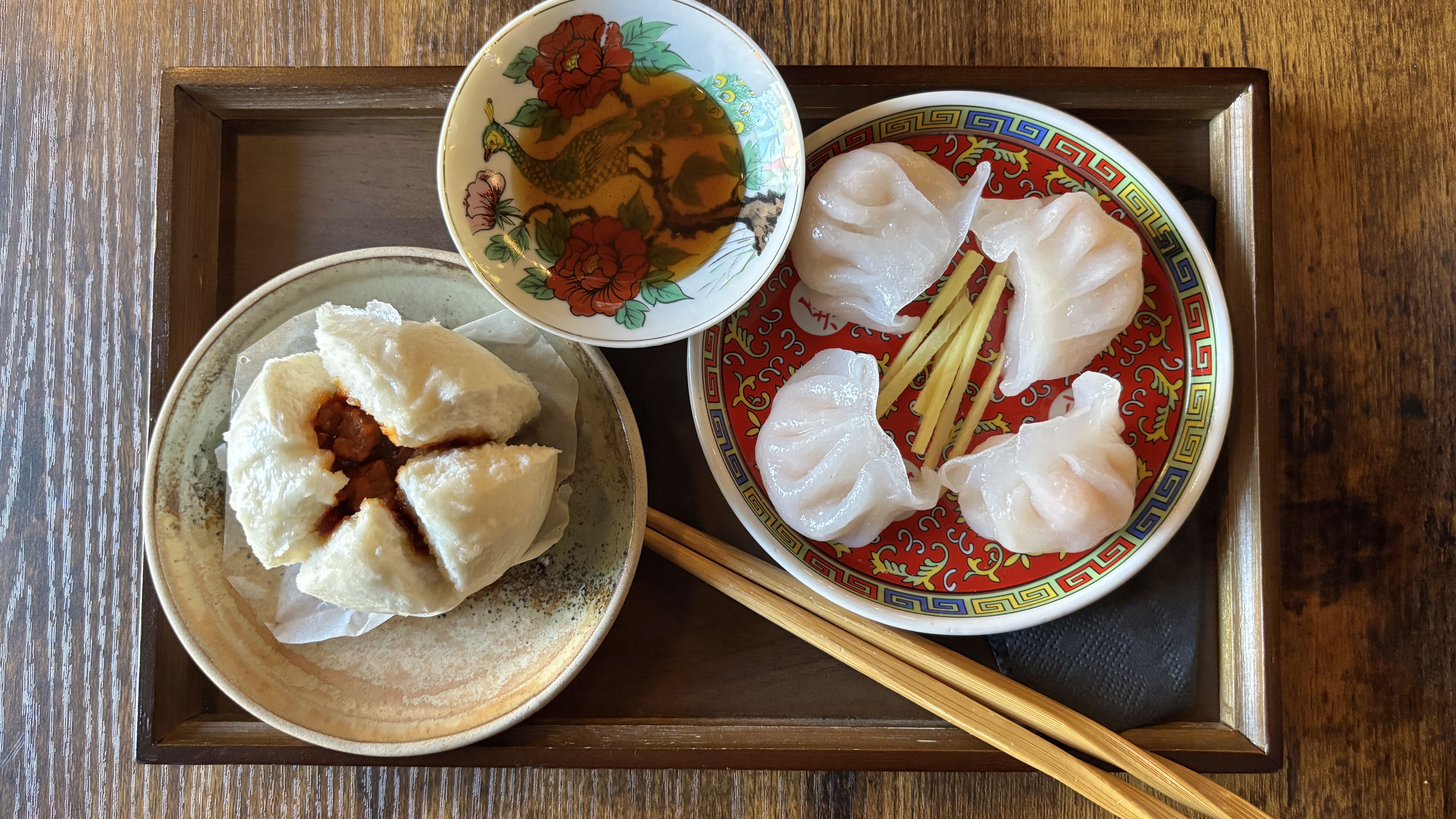 Top-down view of a dim sum tray: white steamed buns with filling on a plate, translucent dumplings on a red and gold plate, dipping sauce in a decorative floral bowl, with chopsticks beside.
