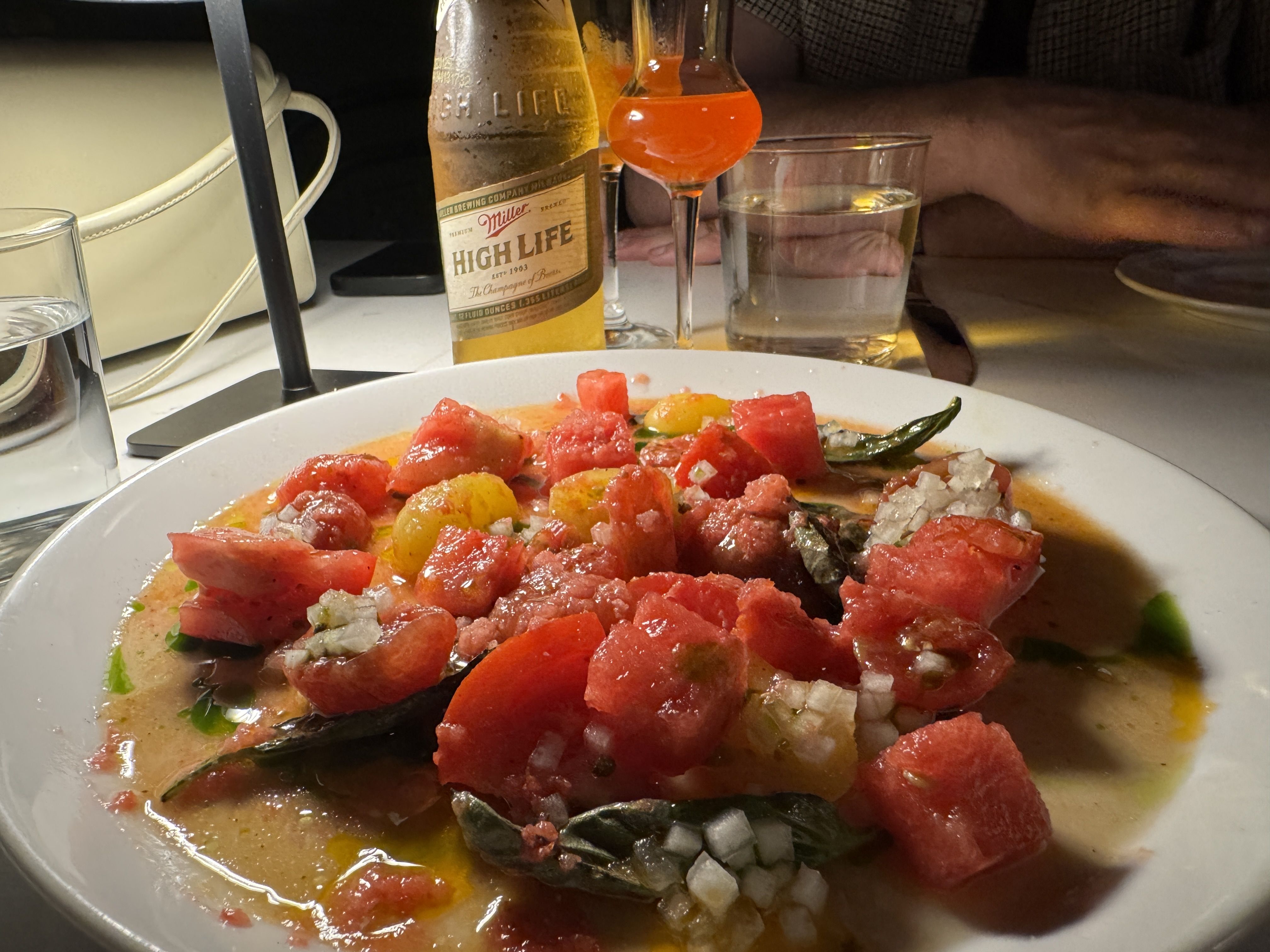 White plate with a salad of watermelon cubes, diced onions, basil leaves, and yellow cherry tomatoes in dressing. In background, a bottle of Miller High Life beer, a glass of water, and a cocktail.