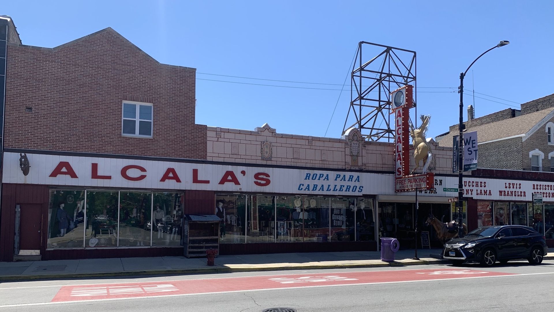 Exterior of Alcalá's store with large red letters and a horse statue on the sign, blue sky, and a black car parked in front. Signs advertise men's clothing brands.