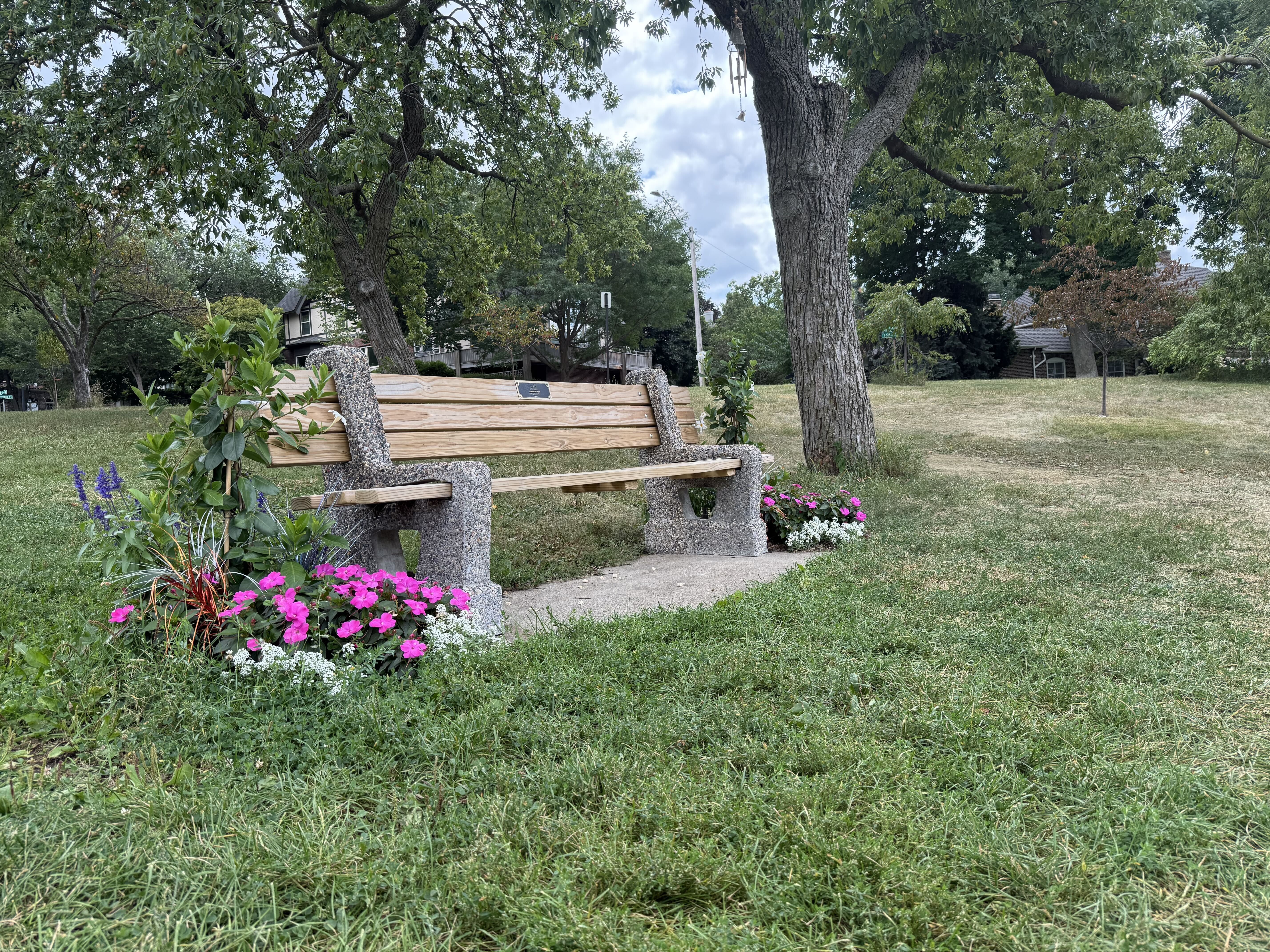 A wooden bench with stone supports sits in a grassy park beneath large trees. Colorful flowers bloom in the foreground; houses and a cloudy sky visible in the background.