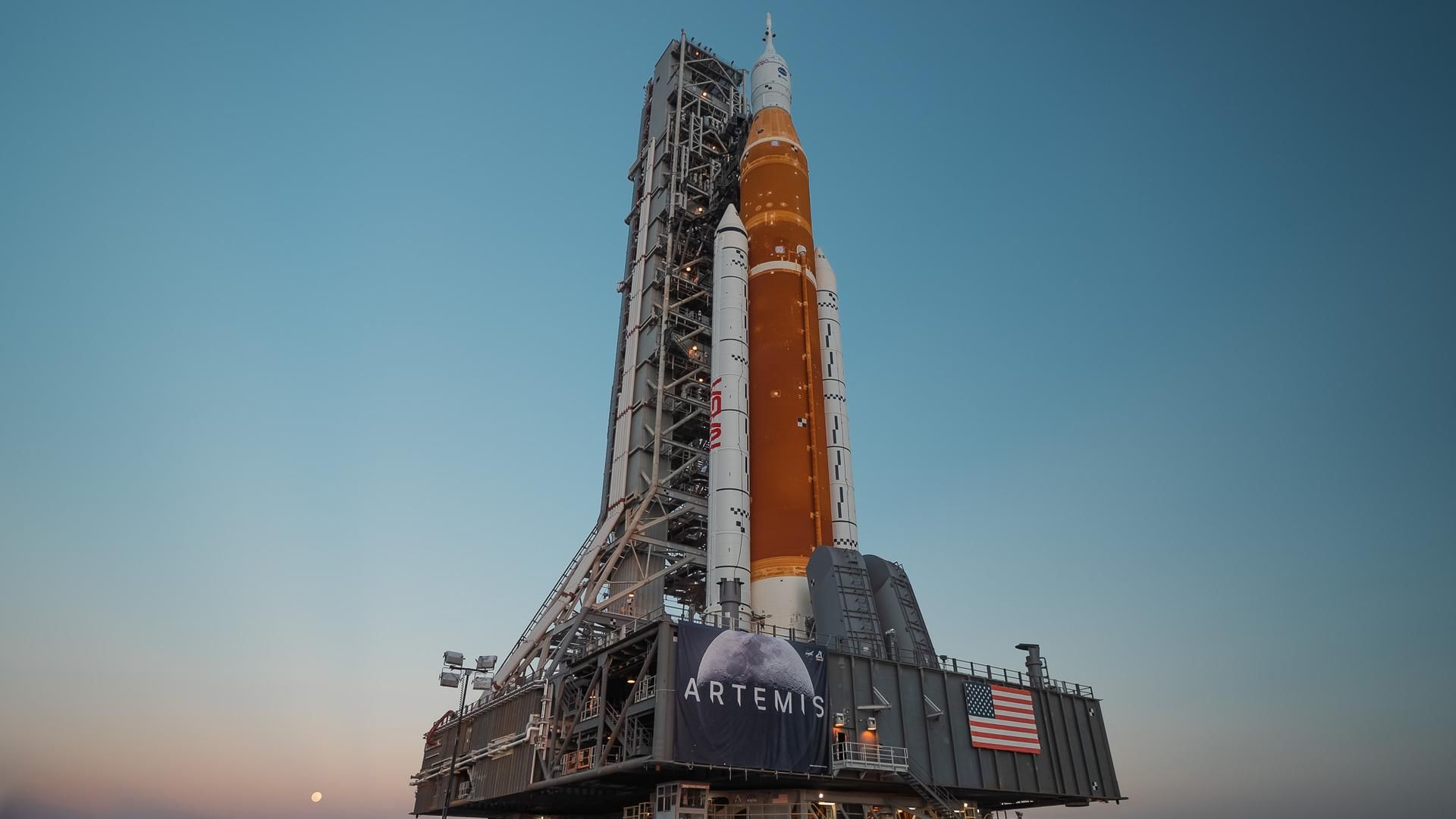 An orange and white rocket stands on a mobile pad against a blue sky