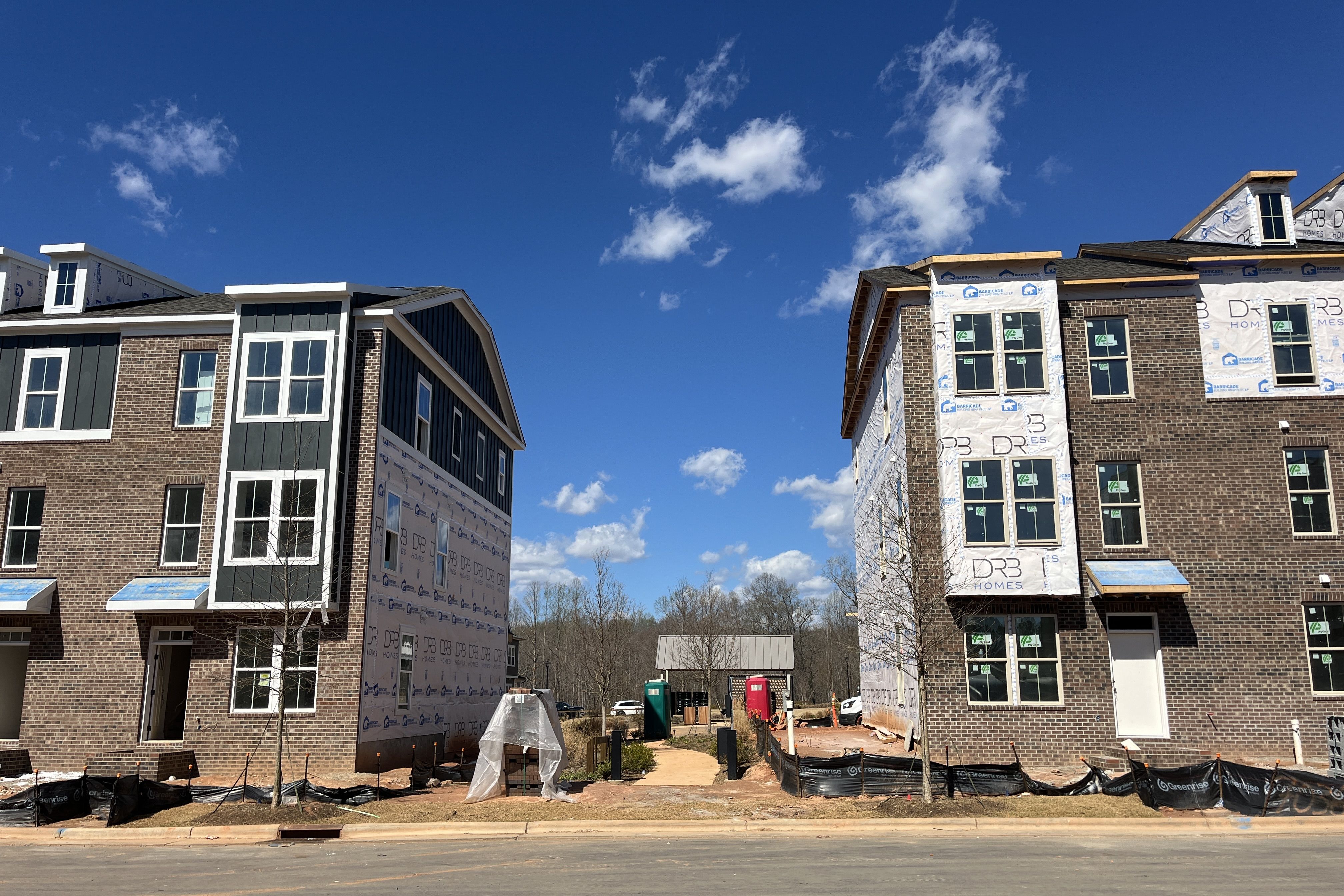 Under-construction row of two three-story brick townhomes flanking a dirt path; left has dark siding and large windows, right is wrapped in white construction wrap under a bright blue sky.
