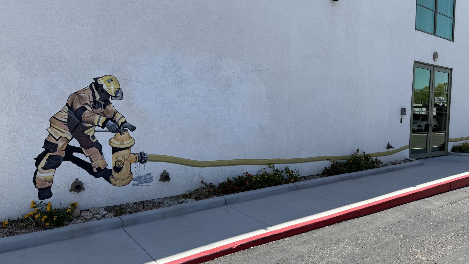 A white office building wall with a painting of a firefighter kneeling next to a fire hydrant, with the hose running along the bottom part of the wall. 