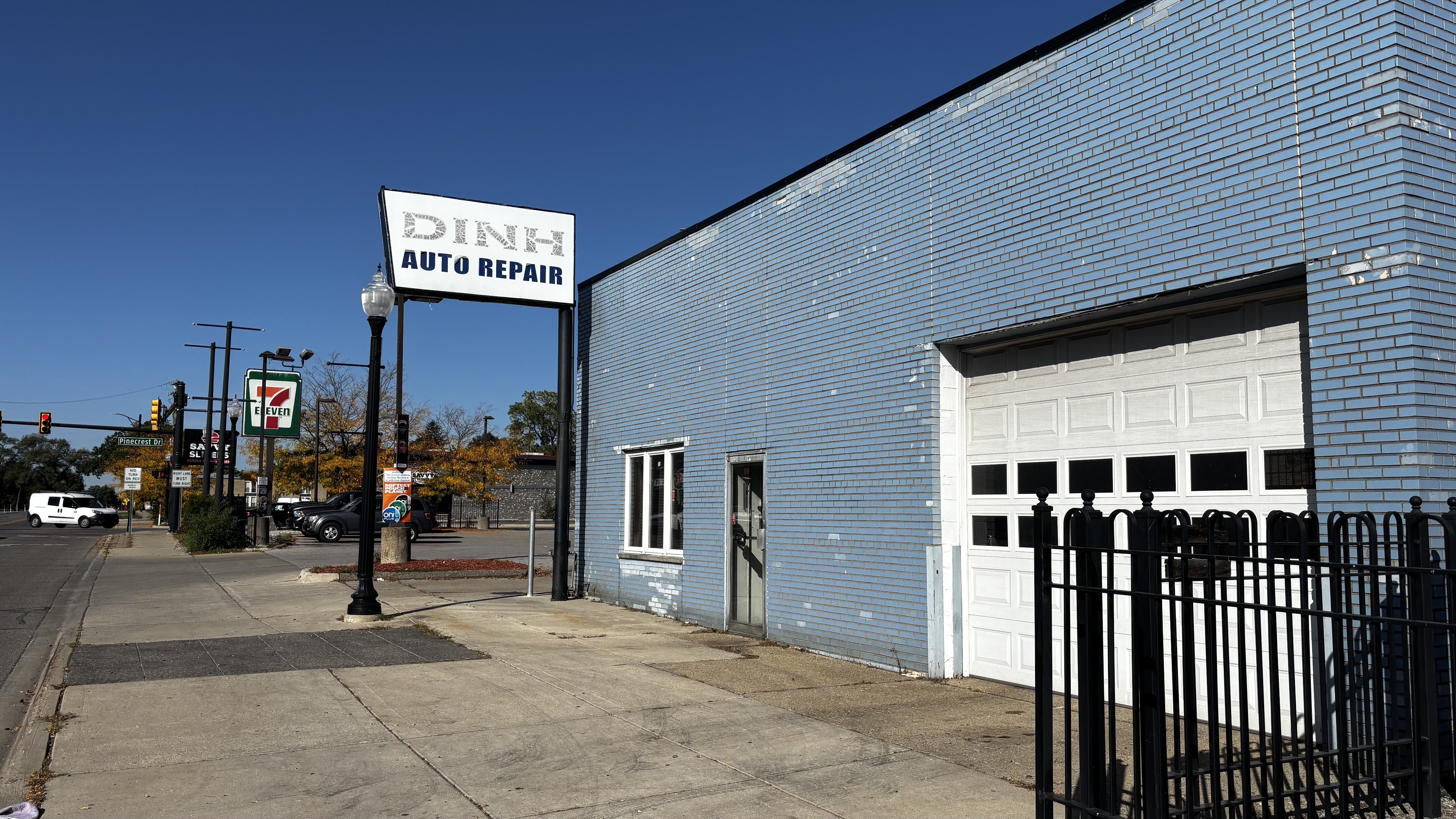 Sunny street scene with a pale blue brick auto repair shop, a large white garage door, a weathered sign reading DINK- AUTO REPAIR, a lamppost, 7-Eleven sign, parked cars, and a clear blue sky.