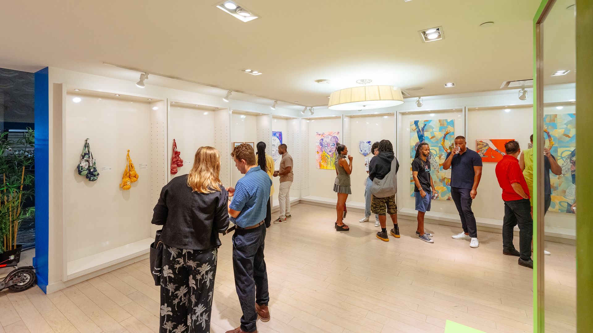 Interior of an art gallery with white walls, hanging artworks and colorful paintings. Group of people chat and observe pieces under ceiling spotlights and a large circular pendant light.