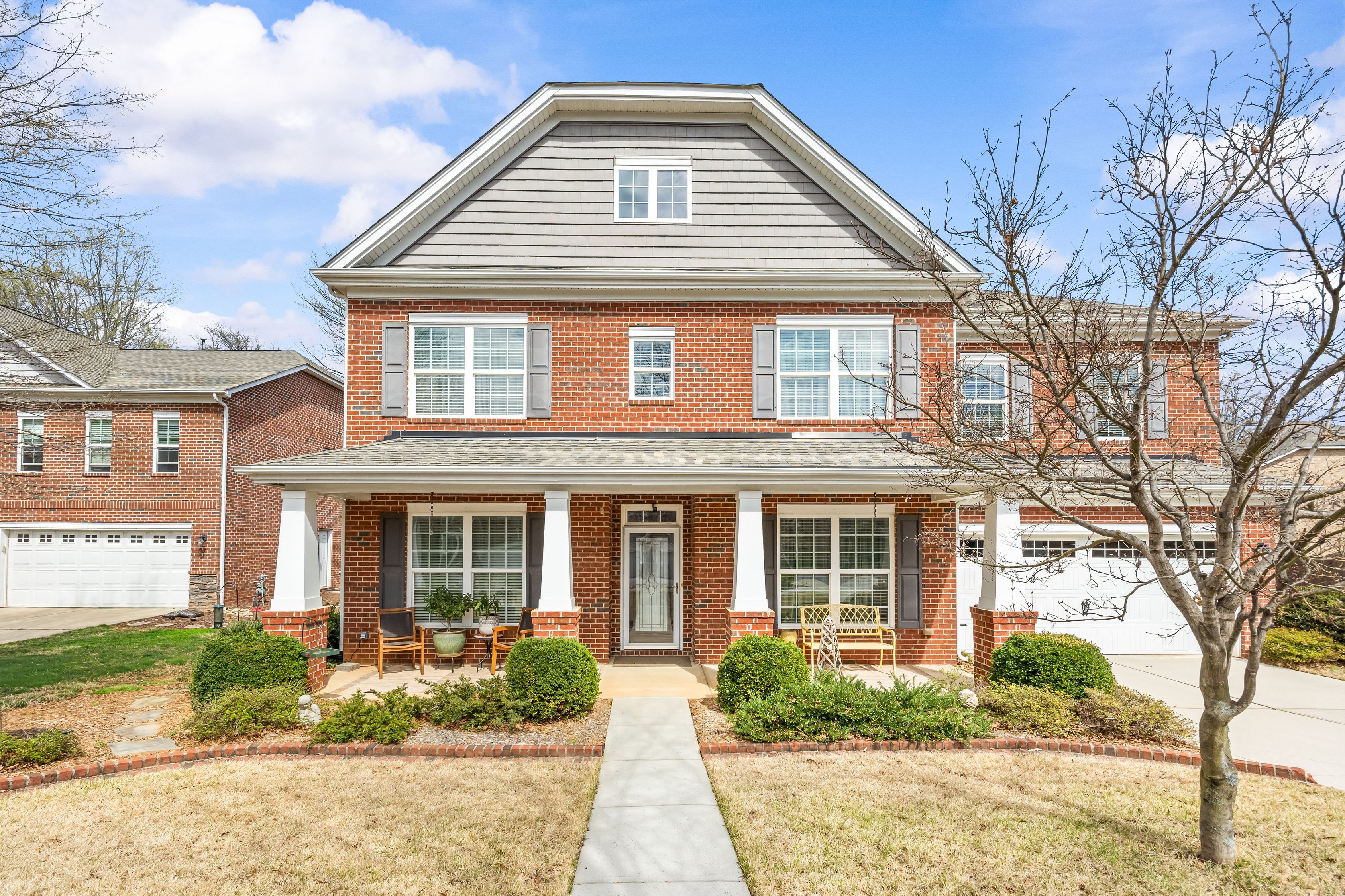 Two-story red brick house with white trim and gray upper siding, a covered porch with white columns, front door, and right-side garage. A leafless tree stands in the yard beneath a blue sky.