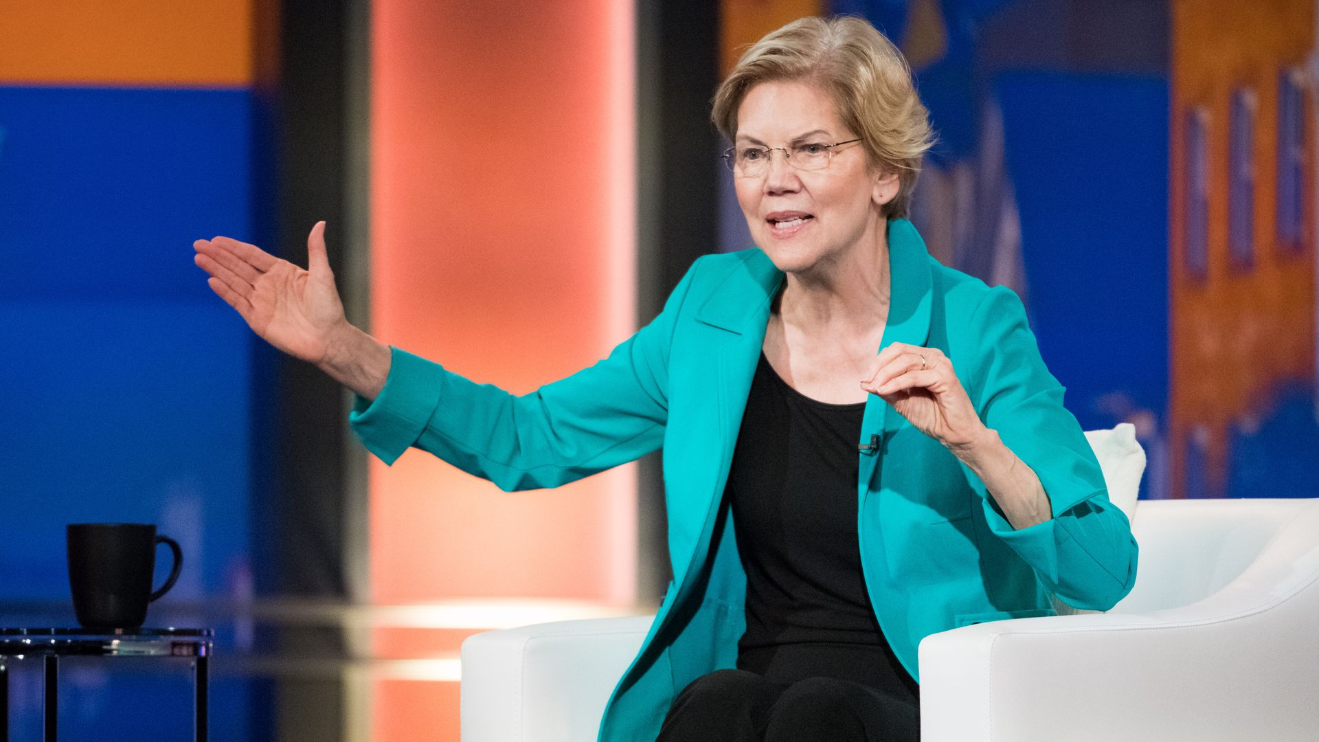 In this image, Warren sits on a white sofa and speaks while gesturing.