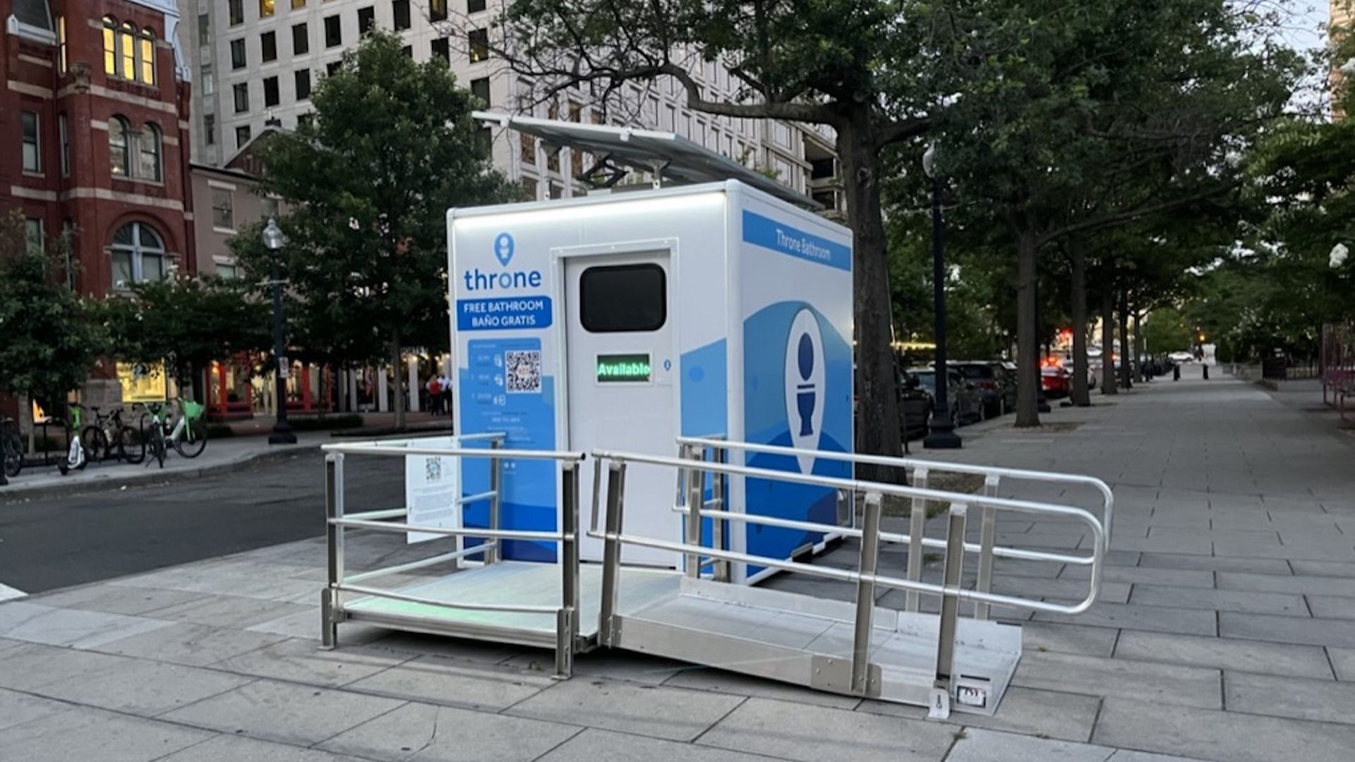 White and blue portable public bathroom labeled "throne" with "FREE BATHROOM BAÑO GRATIS" on a city sidewalk, with an accessibility ramp and trees and buildings in the background.