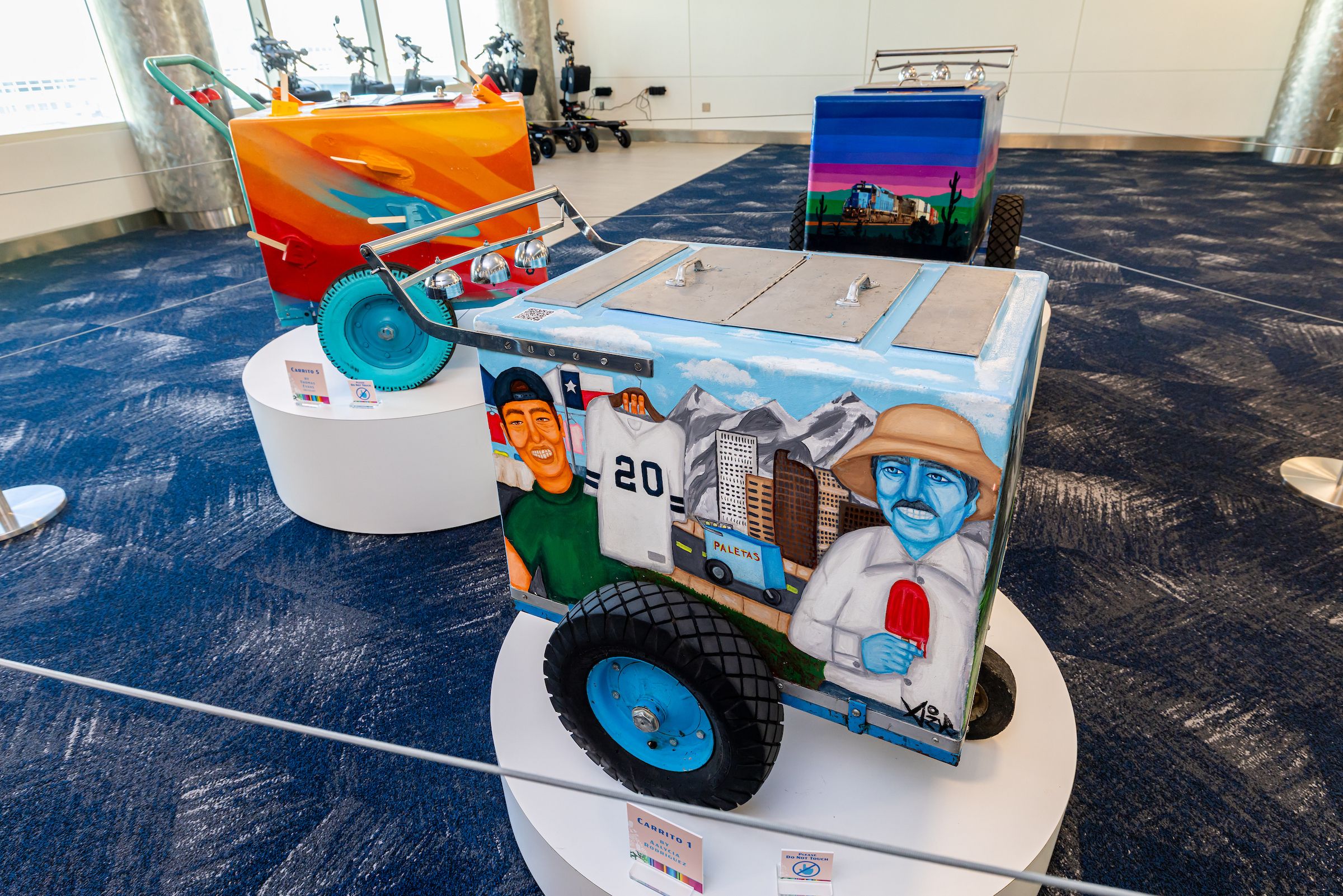 Colorfully painted ice cream carts on display indoors, featuring murals of people, cityscapes, and landscapes with mountains, under bright lighting on round white platforms.