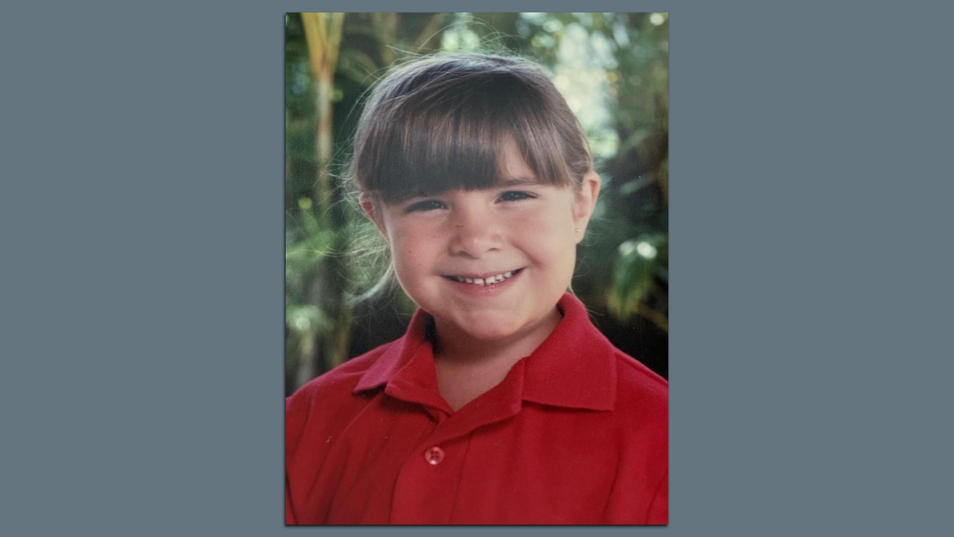 Smiling young girl with brown hair and bangs, wearing a red polo shirt, outdoors with green foliage in a softly blurred background.