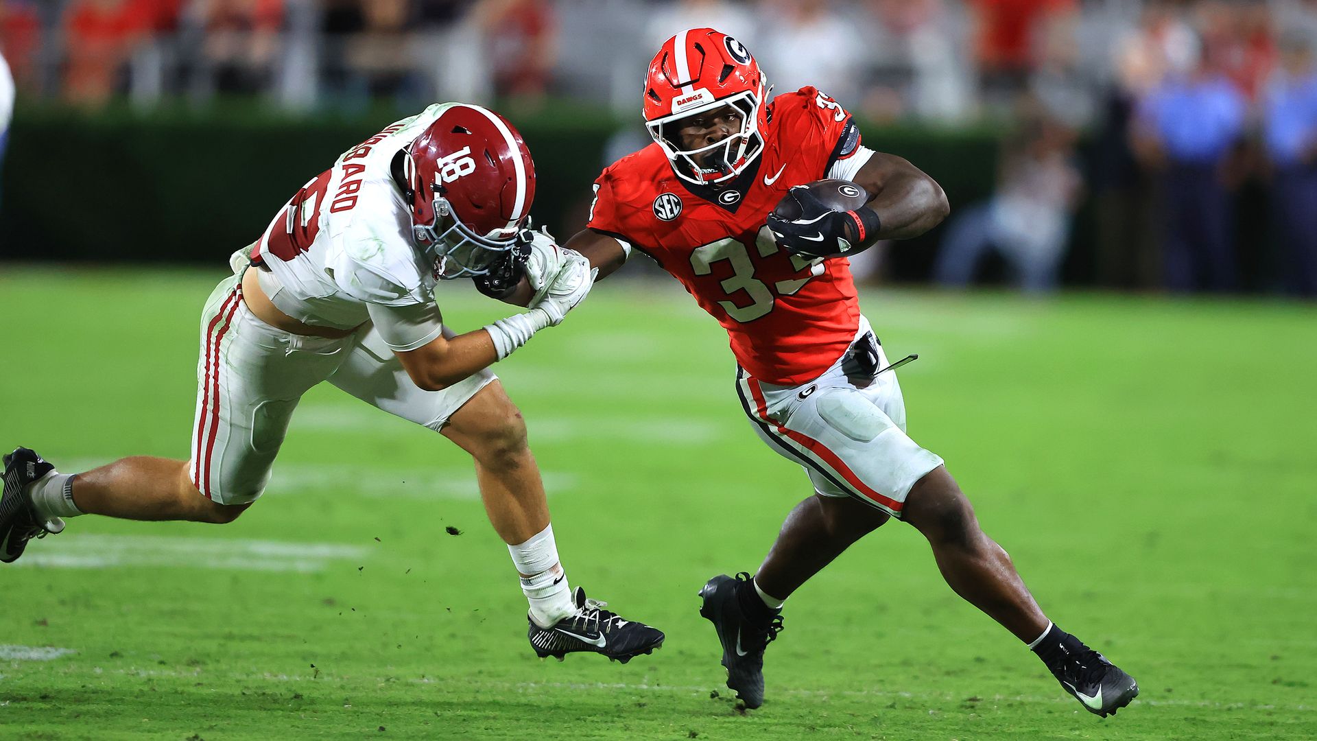 A football player wearing a red Georgia Bulldogs jersey and helmet attempts to hold back a defensive player as he runs the ball on a field.