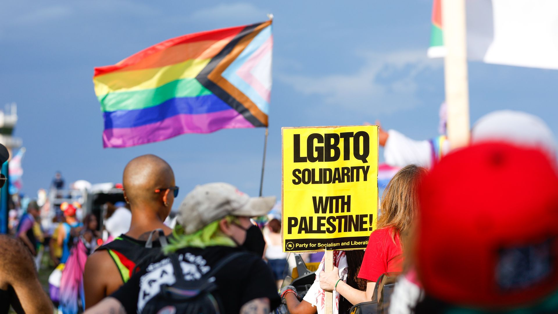 A Progress Pride flag and a yellow sign that reads in black text "LGBTQ SOLIDARITY WITH PALESTINE!" hoisted above a group of people. 