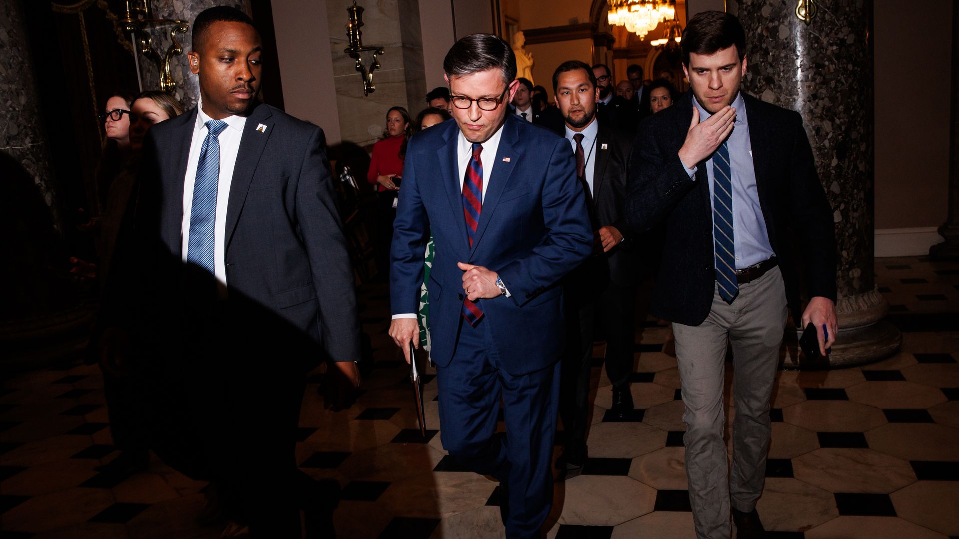 Group of men in dark suits walk through a marble, checkered hallway. Center: man in a blue suit with a red striped tie and glasses, holding a folder; others follow.