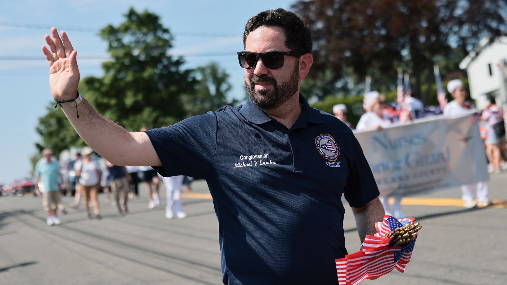 A man wearing dark sunglasses and a navy polo with a circular emblem waves to a parade crowd on a sunny day; spectators and a banner with American flags line the street.