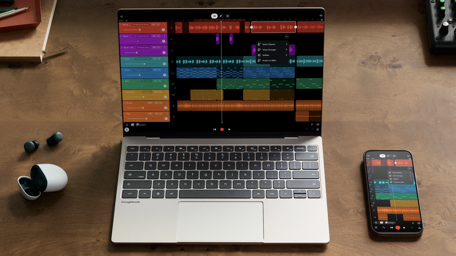 Top-down view of a wooden desk with a silver Chromebook displaying a colorful multi-track music project; a smartphone shows the same app, with wireless earbuds on the left and a small mixer on the right.