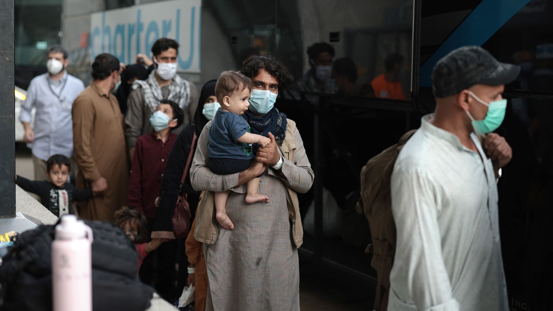 A group of people, including babies and toddlers and adults with blue masks on, walking through an airport with a bus in the background.