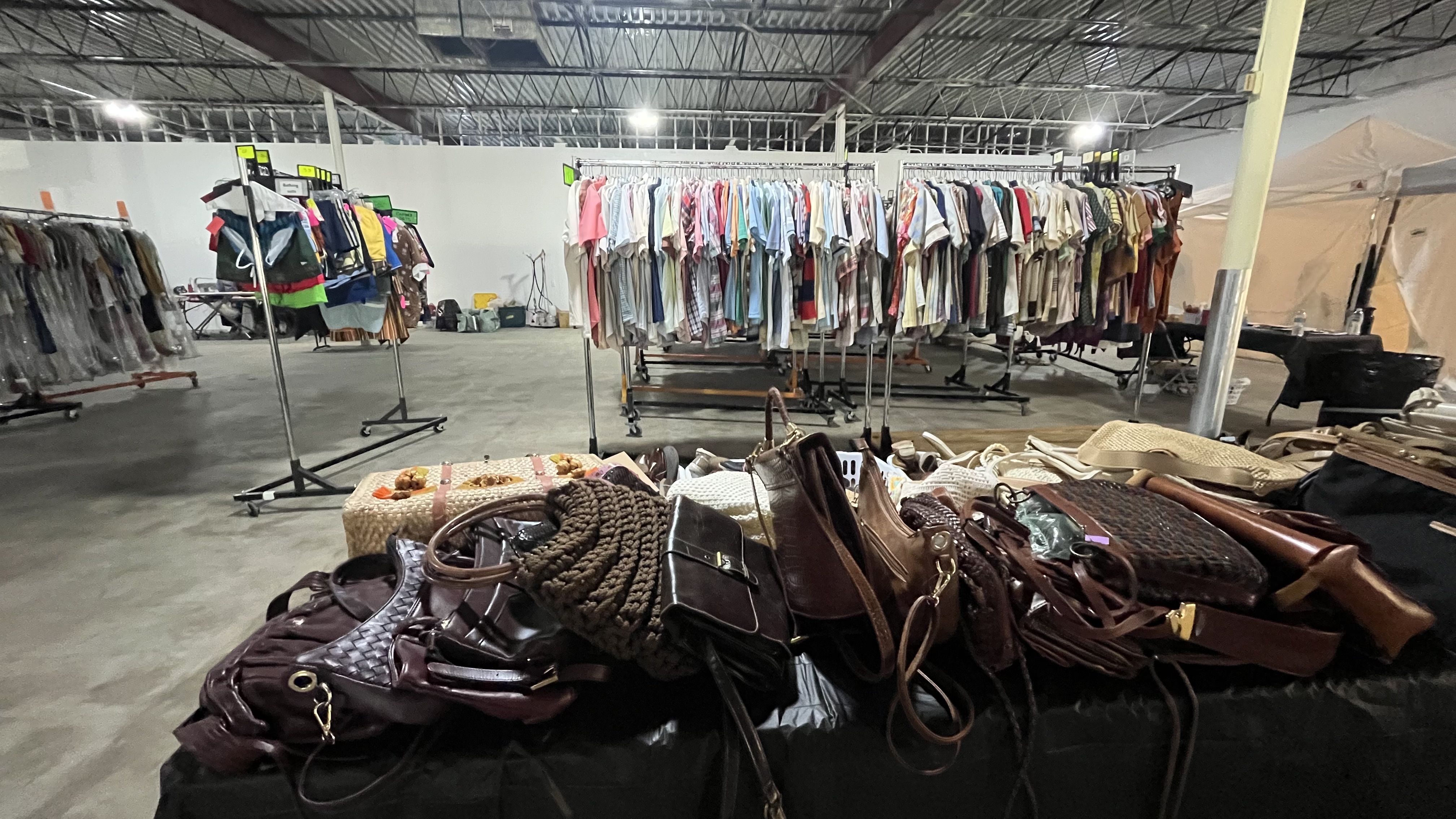 Inside a large warehouse thrift sale: foreground pile of brown handbags on a black table, with racks of colorful shirts on hangers in the background and bright industrial lighting overhead.