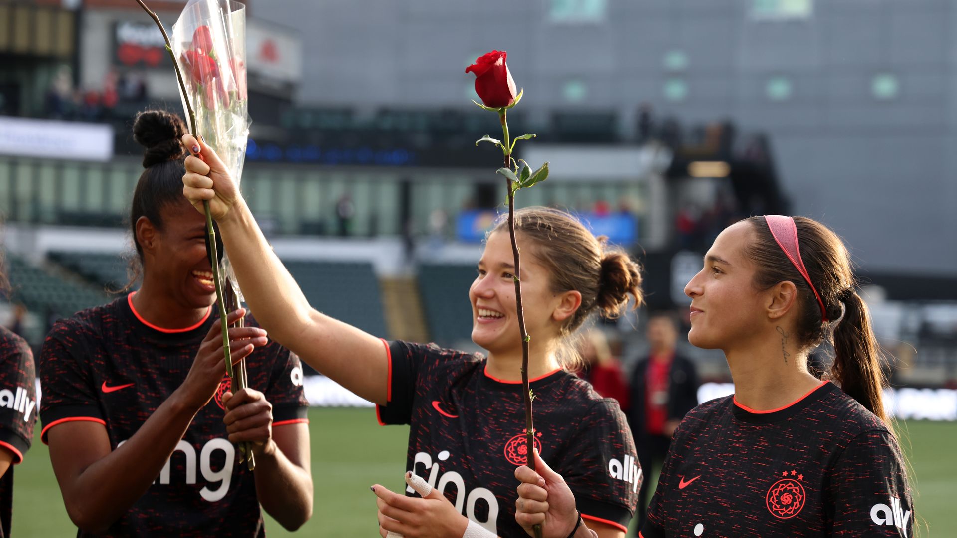Three female soccer players in black jerseys with red accents holding single red roses on a soccer field, smiling and celebrating with a blurred stadium background.