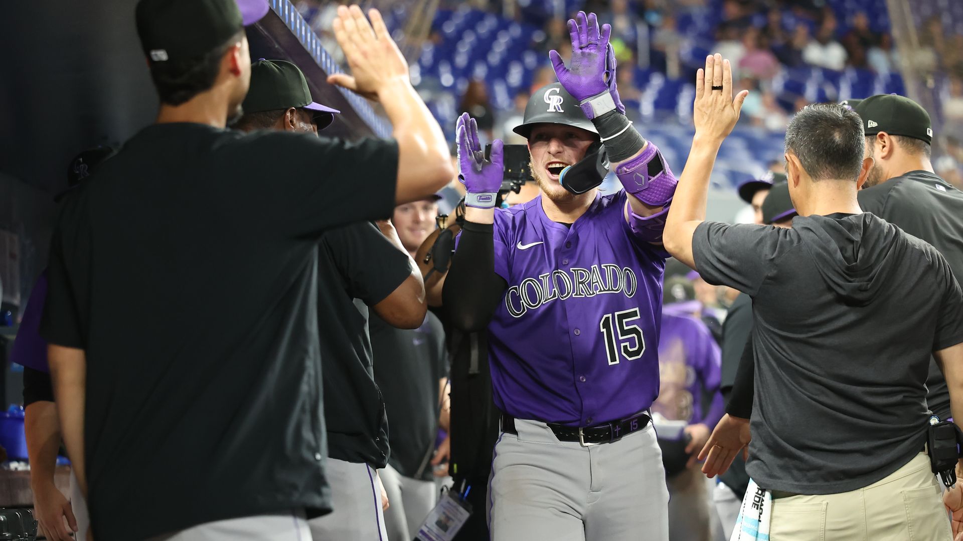 A man raises his hands to high-five other men while wearing a purple baseball uniform and helmet. 