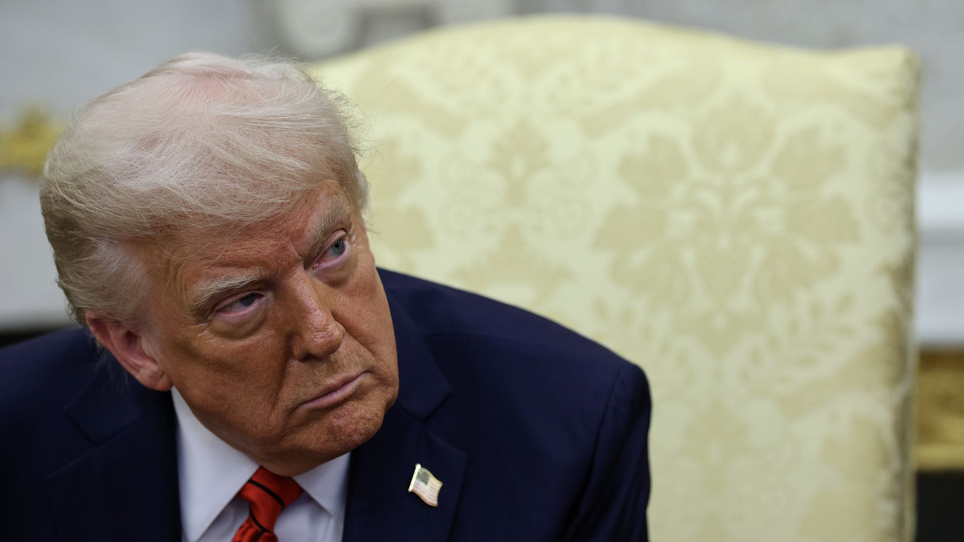 President Trump leans forward in a chair in the Oval Office to listen to a question.