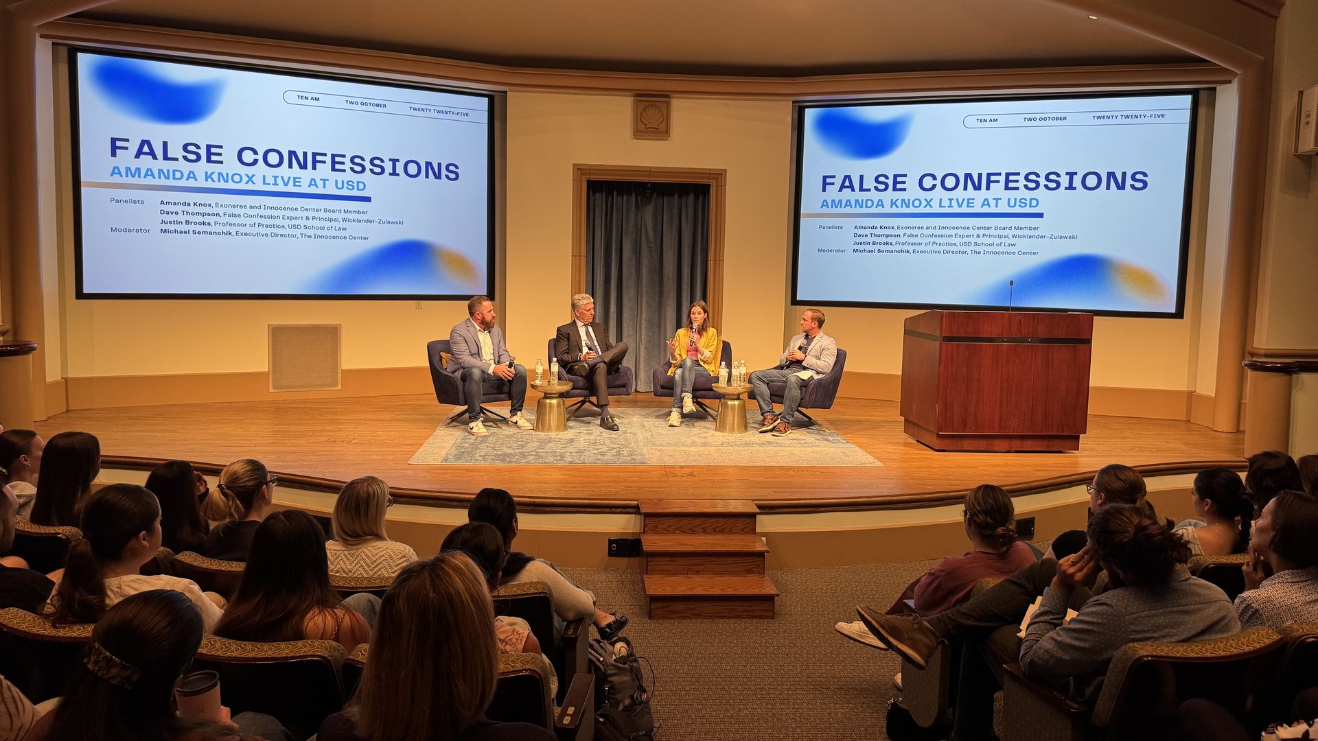Panel discussion titled "False Confessions: Amanda Knox Live at USD" with four speakers on stage, two large screens displaying event title, audience watching in a lecture hall.