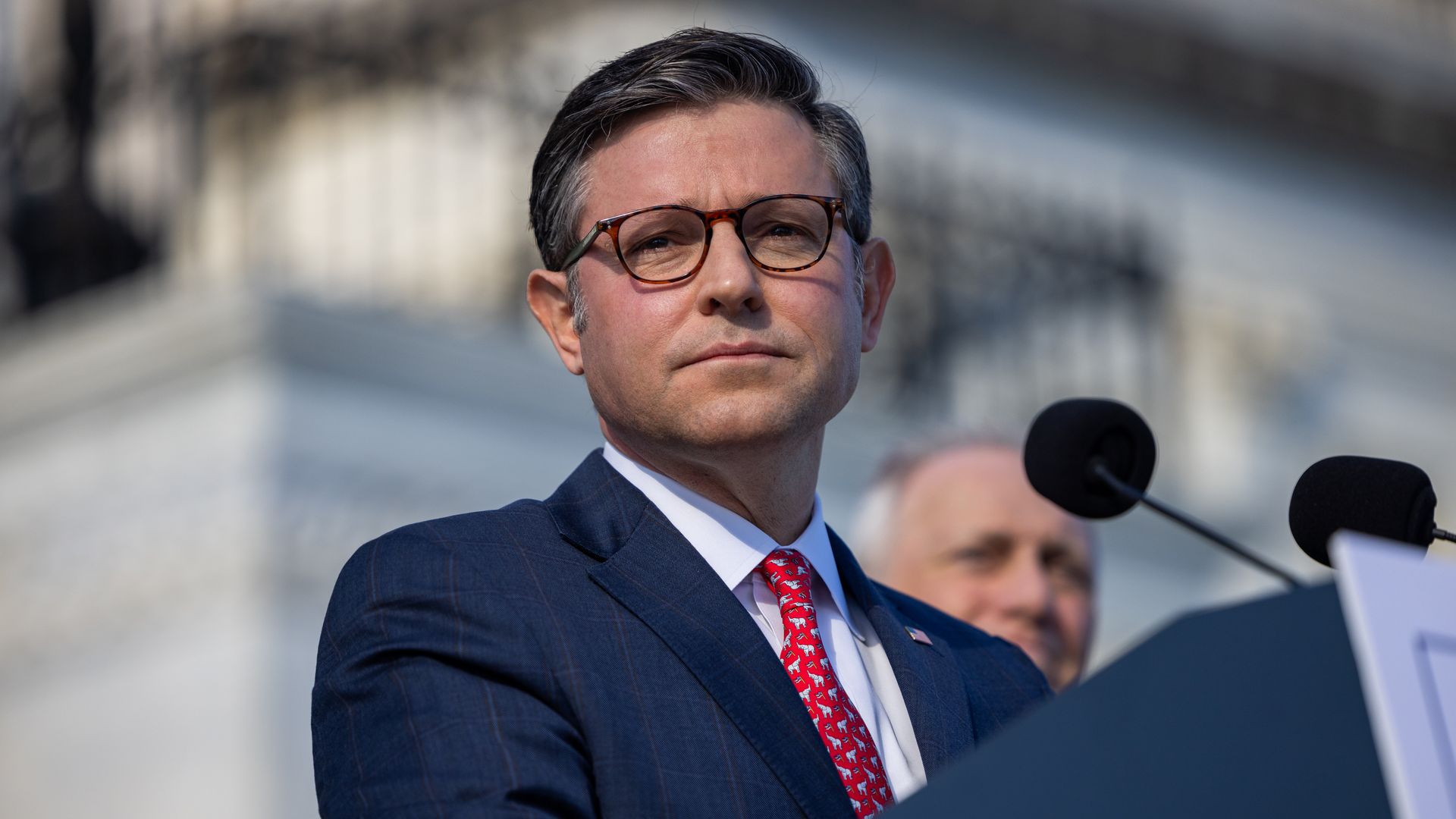 House Speaker Mike Johnson speaking at a podium in front of a marble building.