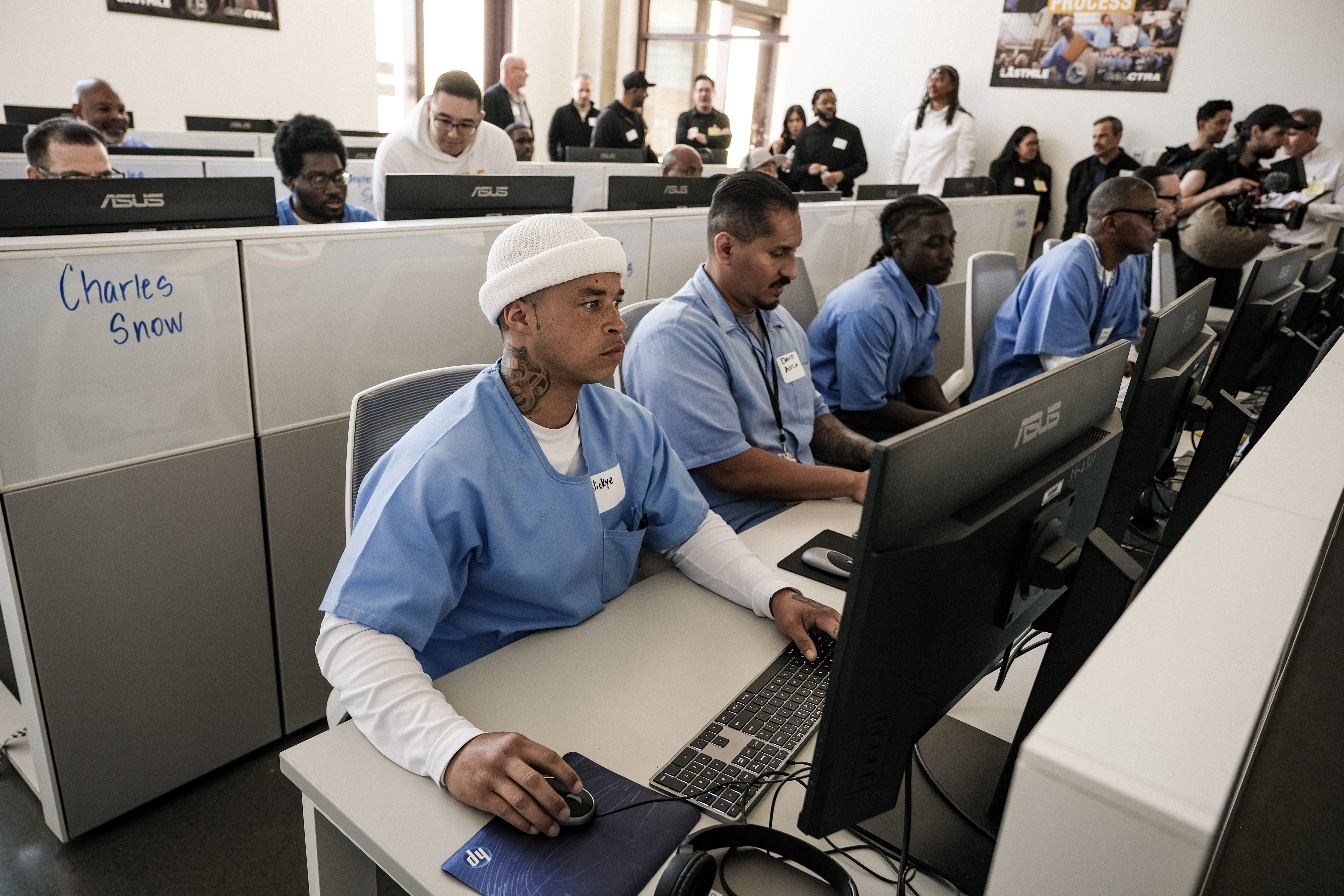 Group of people in a computer lab workshop. In the foreground, a man in a white beanie and blue scrubs sits at an ASUS monitor; others work at desks while attendees stand in back.