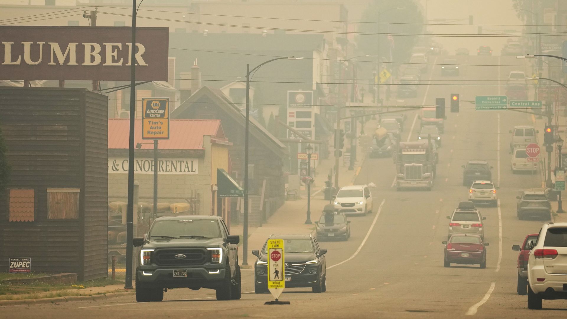 Town street in thick beige haze; a large LUMBER sign on the left; power lines and street lamps cross the road; cars line the boulevard with a STOP sign and a yellow pedestrian sign in the foreground.