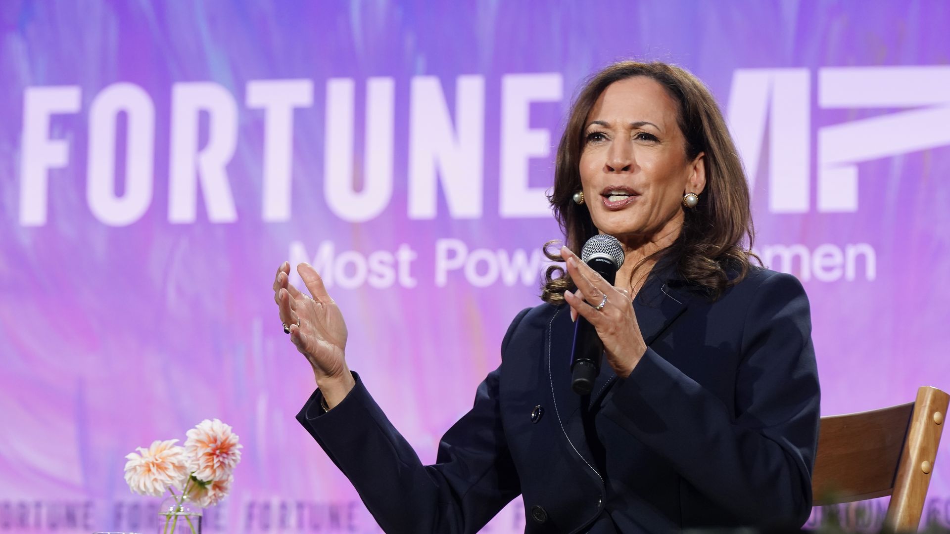 Kamala Harris, dressed in a black suit, speaks into a microphone with a purple and pink background.