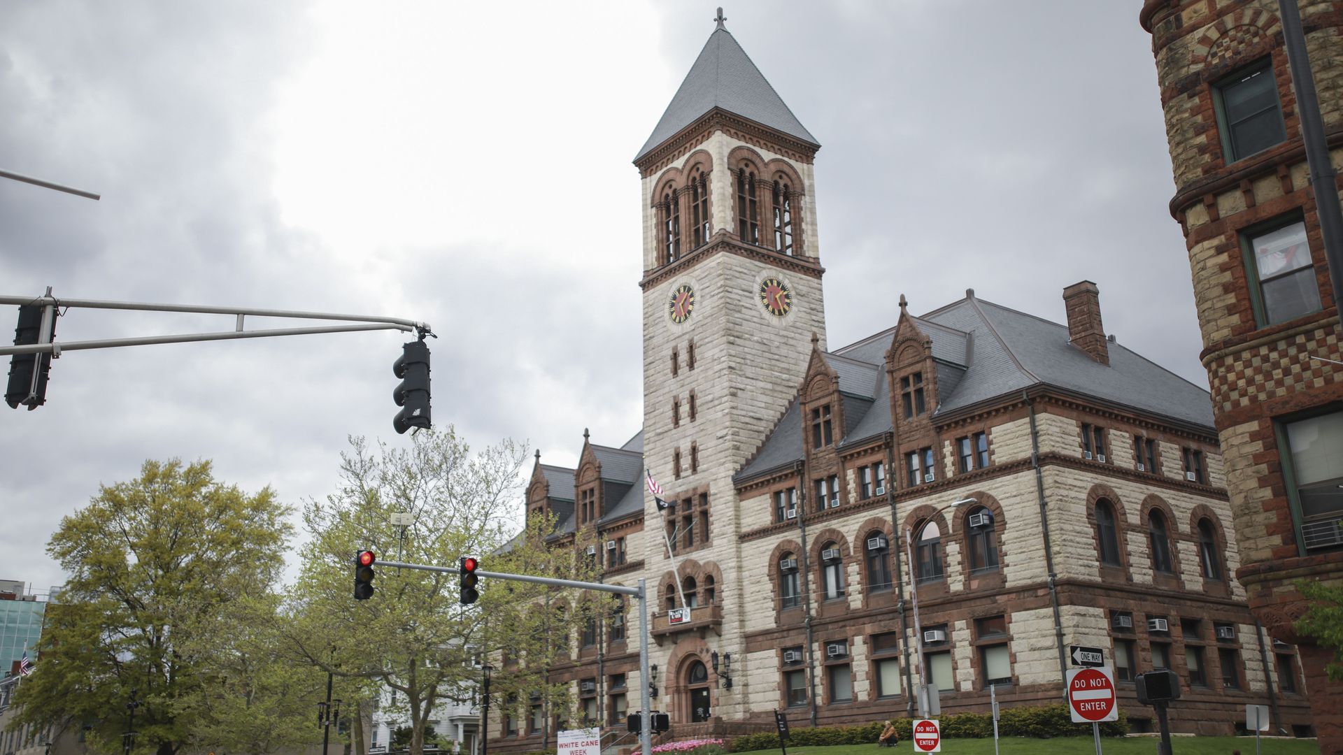 Historic stone building with a tall clock tower, arched windows, and a gray roof under a cloudy sky, surrounded by trees, traffic lights, and no entry signs.