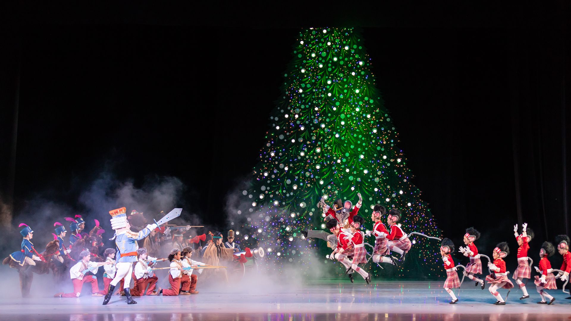 Ballet performers dressed in Nutcracker costumes gathered around a christmas tree on stage.