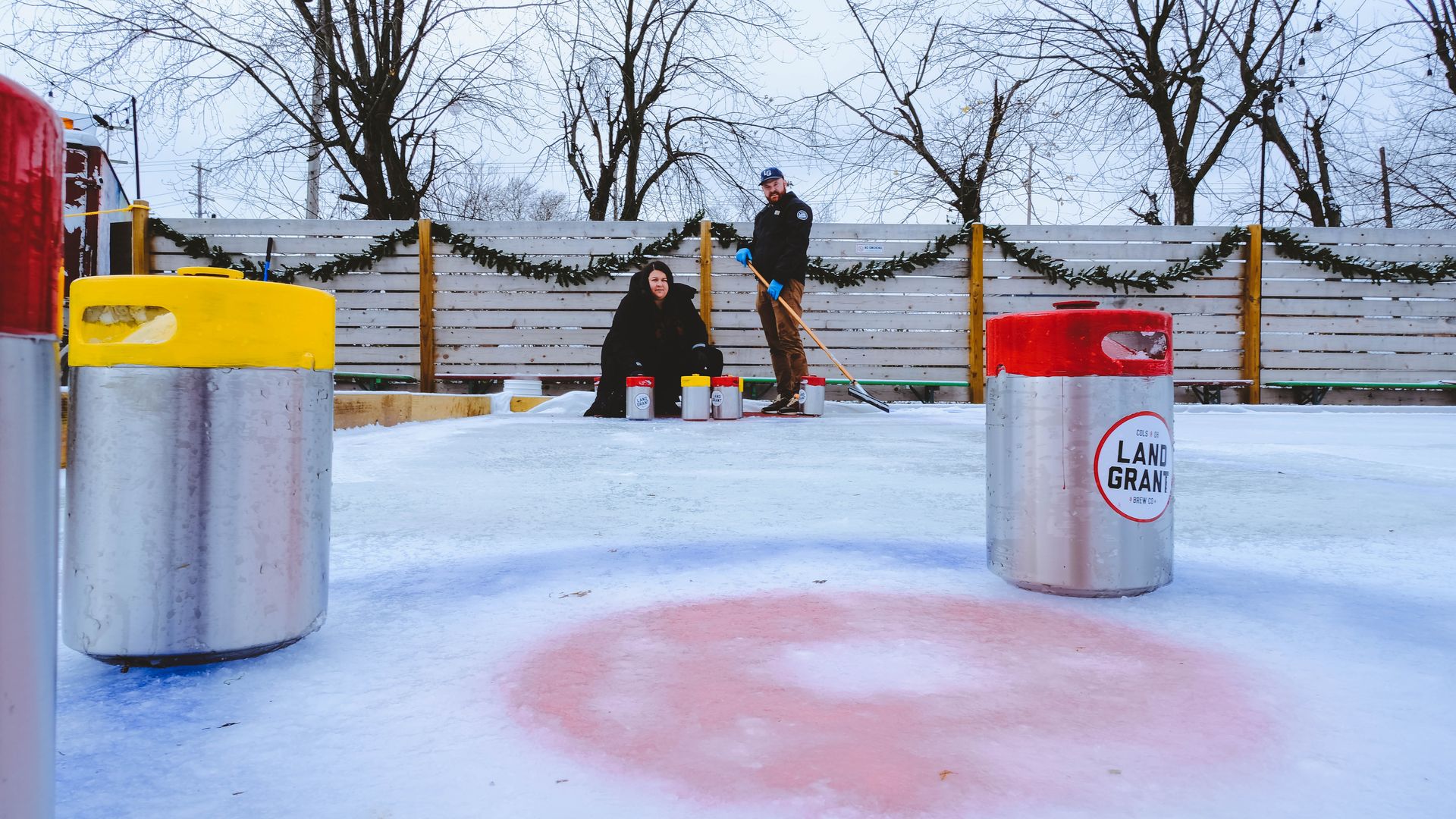 Two people watch beer kegs slide on ice at the end of a curling rink