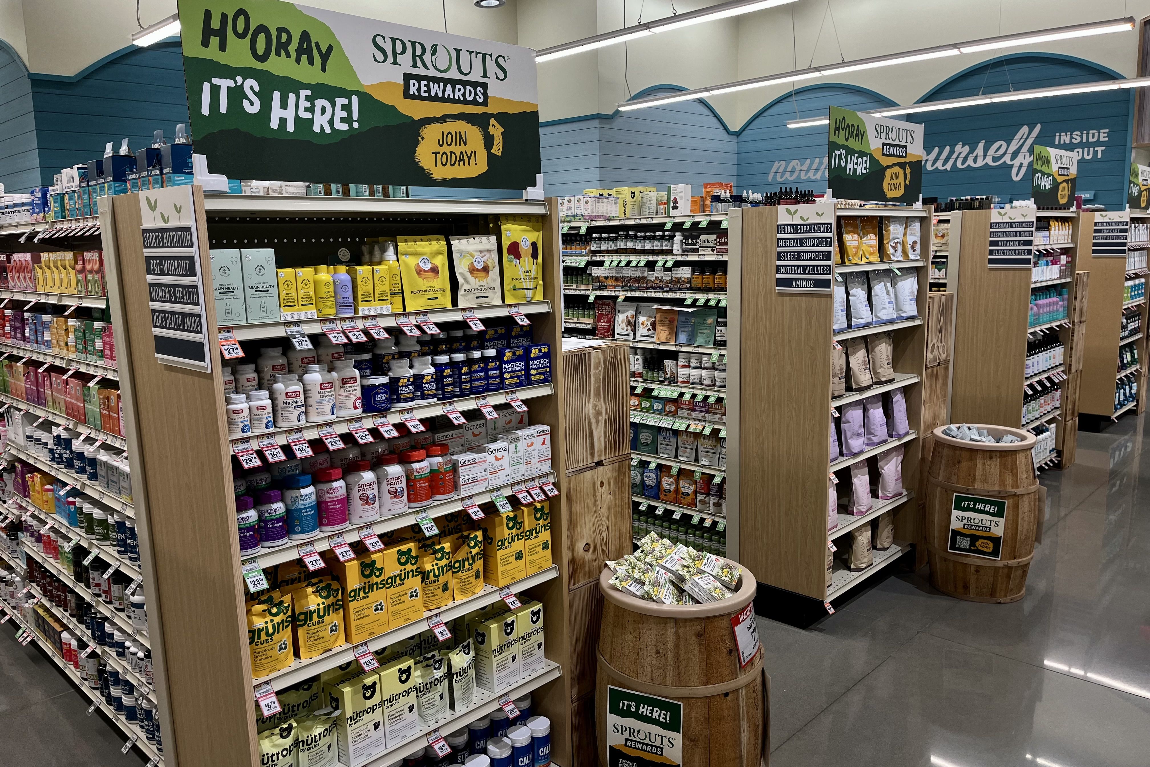 Interior of a Sprouts store showing aisles of health supplements and vitamins with wood shelves and signs promoting Sprouts Rewards program in green and yellow.