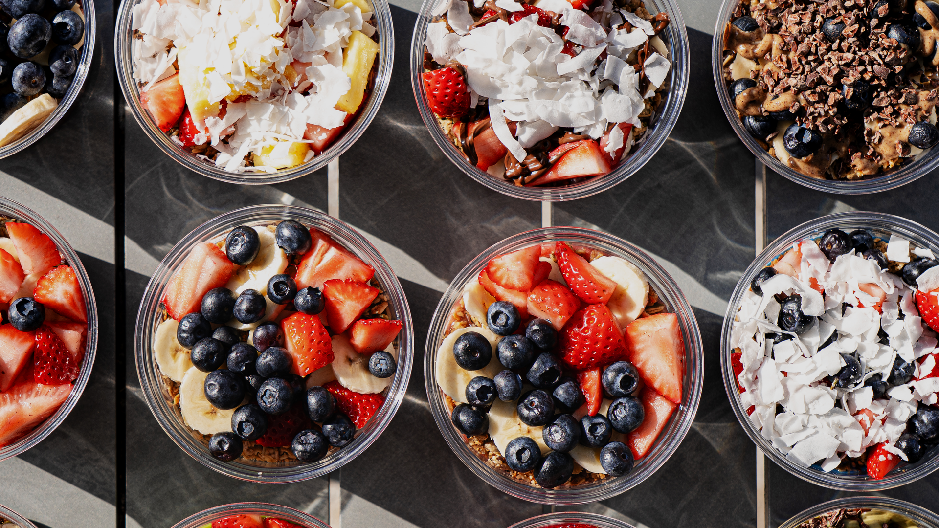 Top-down view of round glass bowls on a dark surface, each filled with yogurt parfaits topped with blueberries, strawberries, banana slices, granola, and coconut flakes.
