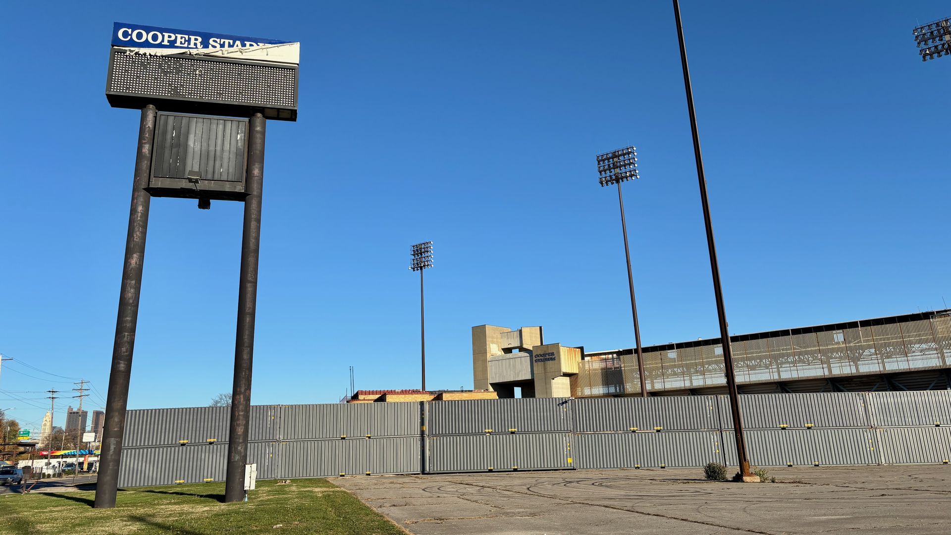 Empty parking lot with a tall, ripped sign that reads "COOPER STA" and a stadium structure behind a gray fence under a clear blue sky.