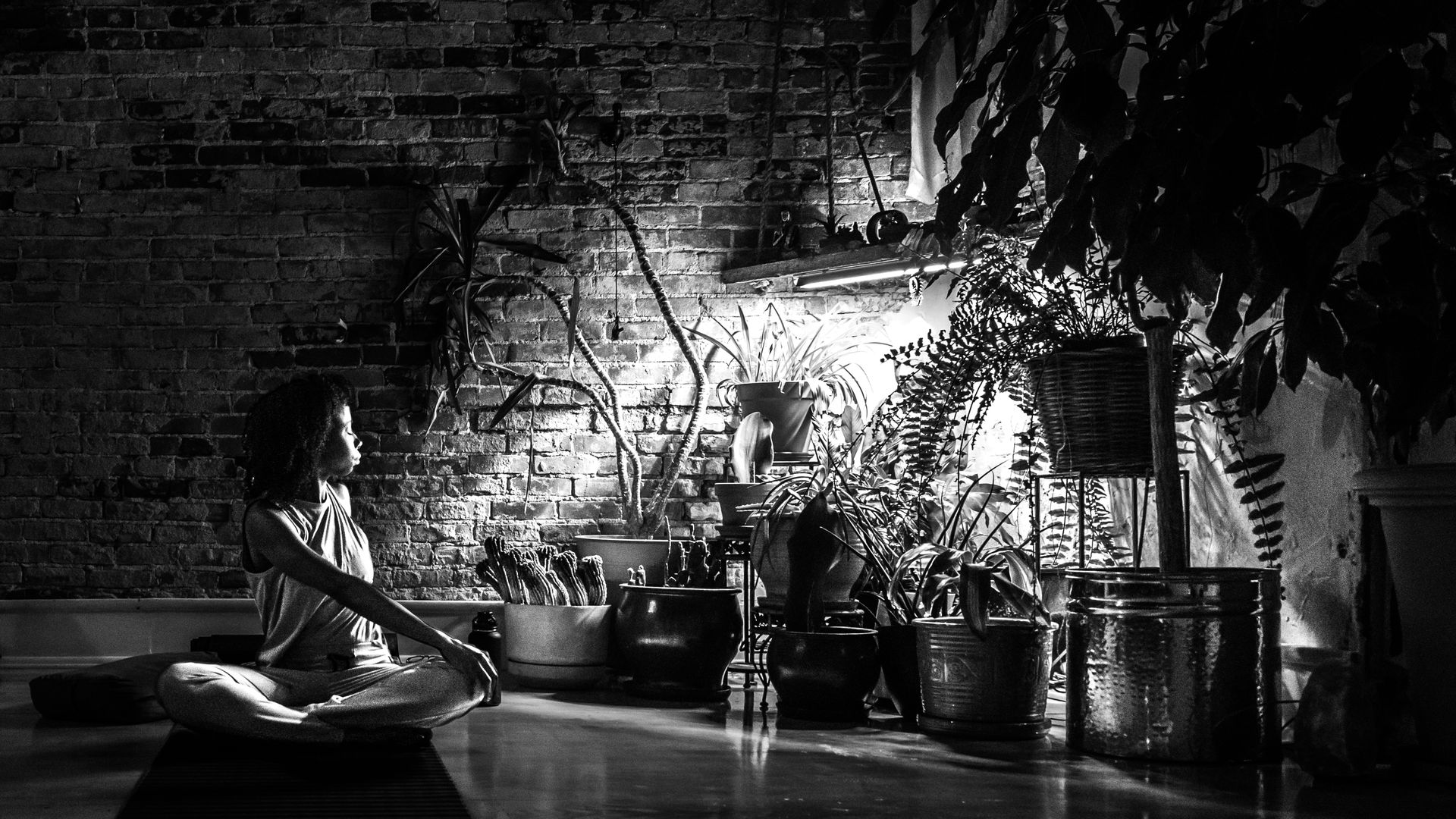 A black and white photo of a woman stretching on her yoga mat surrounded by plants. 