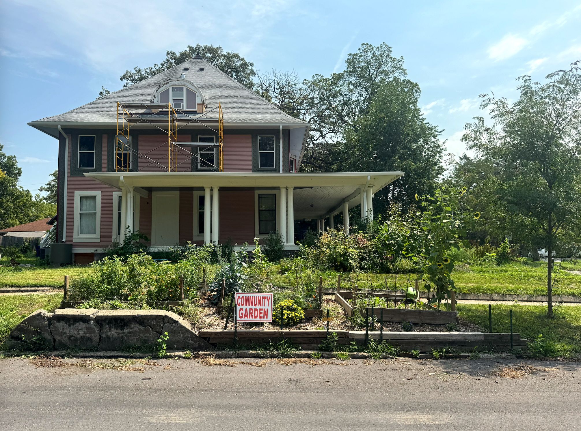 Two-story pink house with a gray shingled roof and yellow scaffolding on the upper level. White-columned porch; foreground shows garden plots and a sign reading COMMUNITY GARDEN under a blue sky.