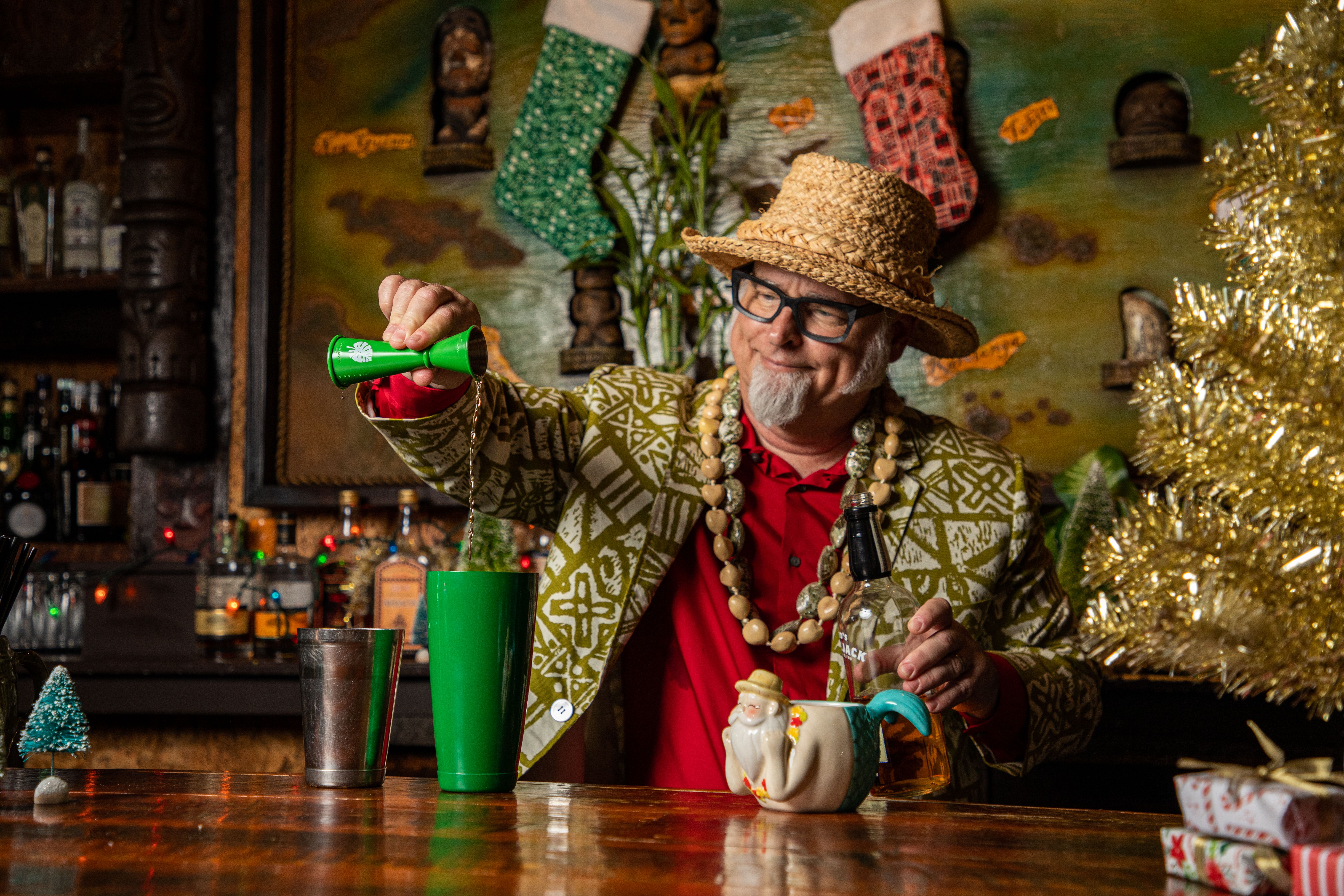 Jeff Berry mixes a cocktail. He stands behind a tiki bar and pours liquid from a green jigger into a green cocktail shaker.