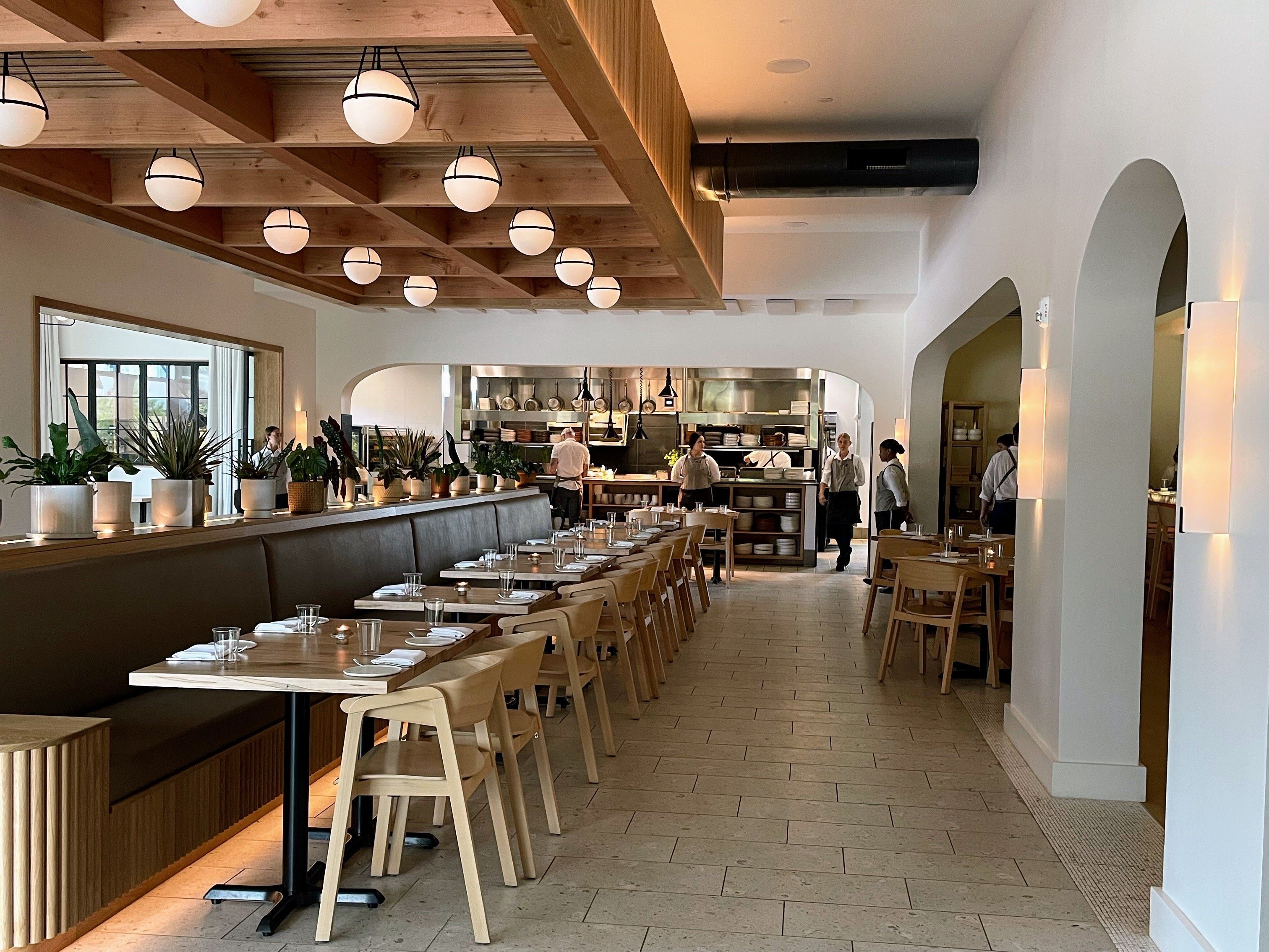 Bright, modern restaurant interior with exposed wooden beams and round globe pendant lights. Long tables set with napkins, potted plants along a counter, and an open kitchen visible in the back.