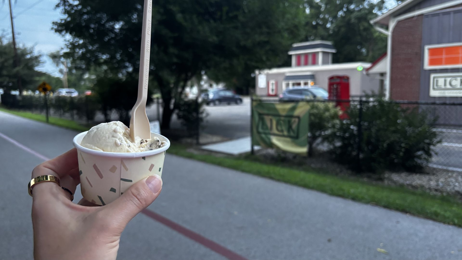 Hand holding a cup of ice cream with a wooden spoon on a path, with trees and a building with a red phone booth in the blurry background during daytime.