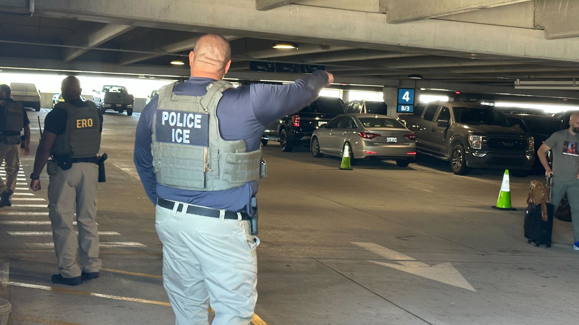 A police officer wearing a vest labeled POLICE ICE points left in a dim parking garage. Another officer with ERO on his vest stands nearby as parked cars and green traffic cones fill the lot.