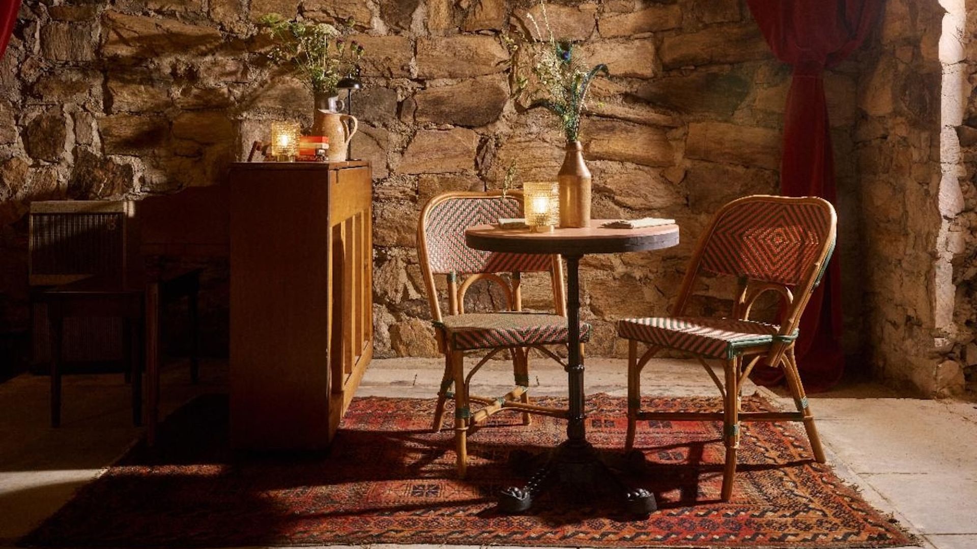 Cozy rustic dining nook with a stone wall, a round table flanked by two wicker chairs, warm candlelight from a bottle vase, a patterned rug, and a red curtain to the right.