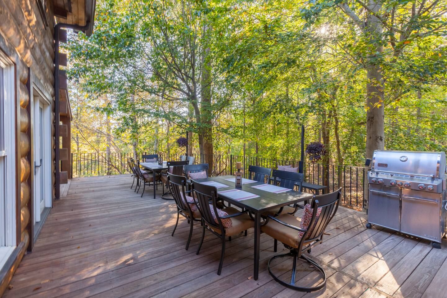 Outdoor wooden deck with black metal dining tables and cushioned chairs, surrounded by green trees, next to a log cabin wall and a silver gas grill under sunlight.