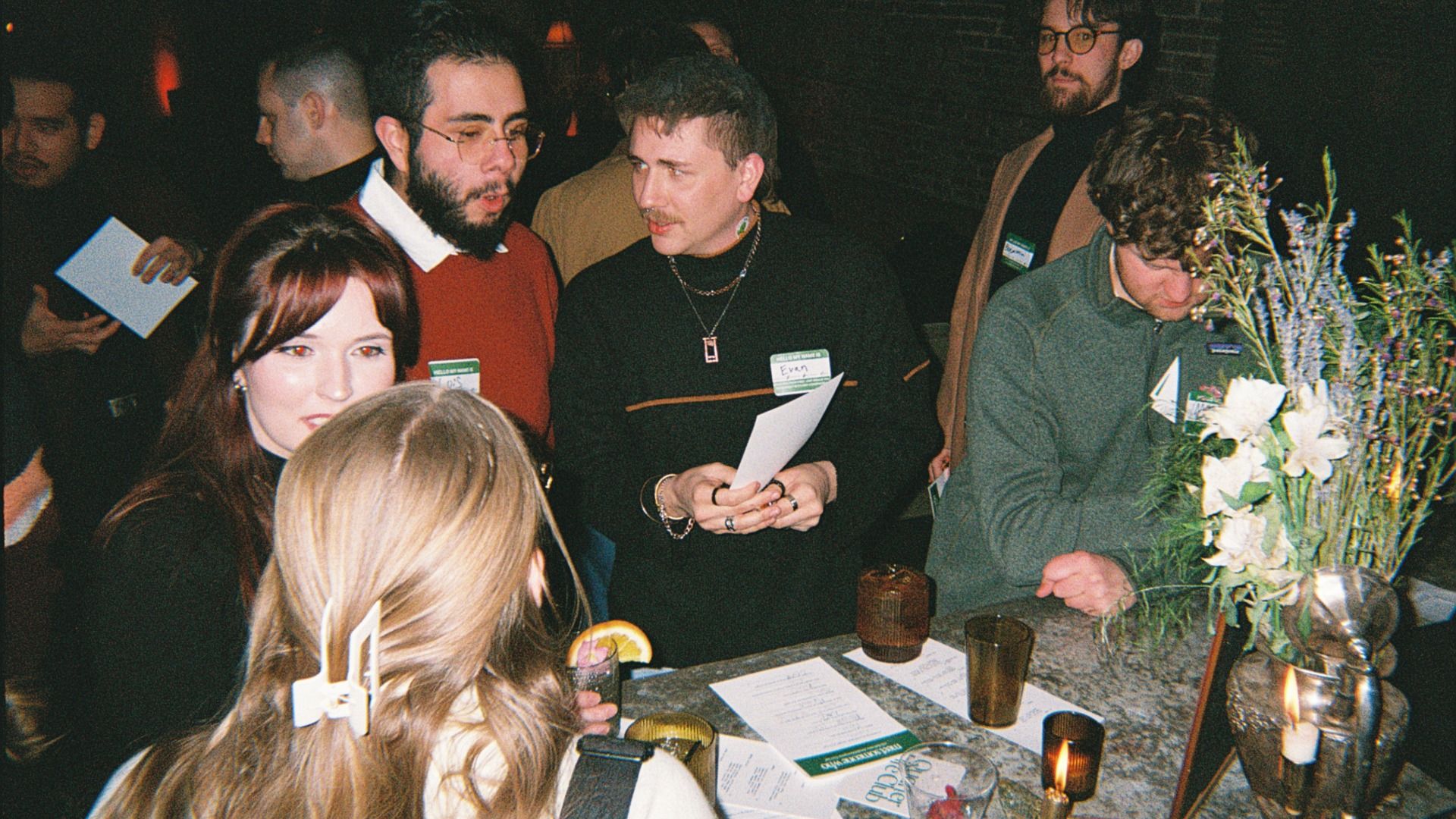 Dimly lit social gathering around a table with papers and drinks; two men in center converse (one in red sweater, the other in black); a bouquet stands at right, a woman in foreground.